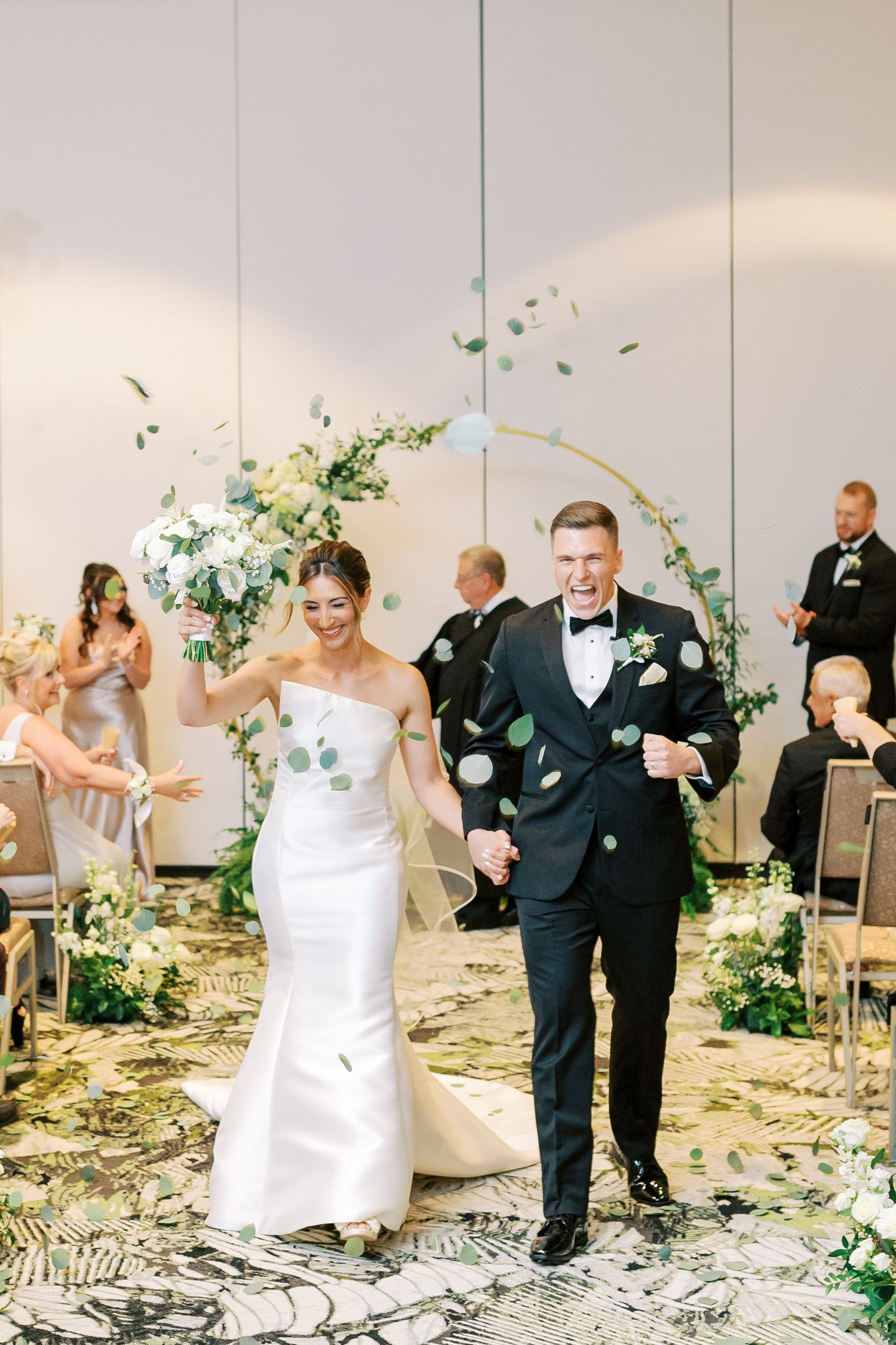Wedding celebration with guests throwing confetti under a floral arch in an elegant indoor setting.