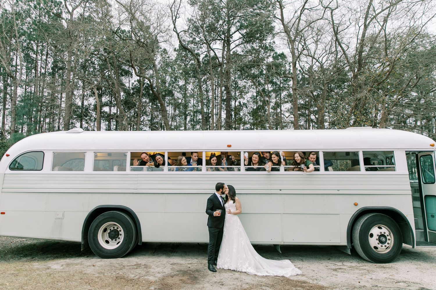 A vintage white bus parked in a wooded area serves as a backdrop for a romantic wedding photo.