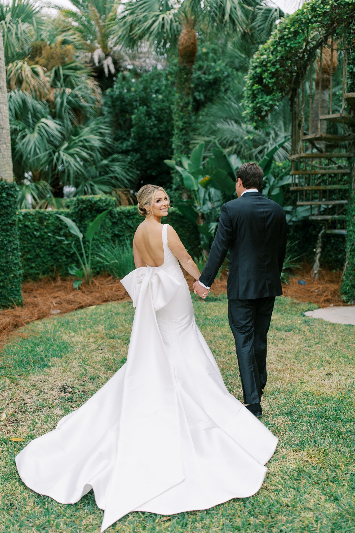 A romantic wedding couple walks through a lush tropical garden with palm trees.