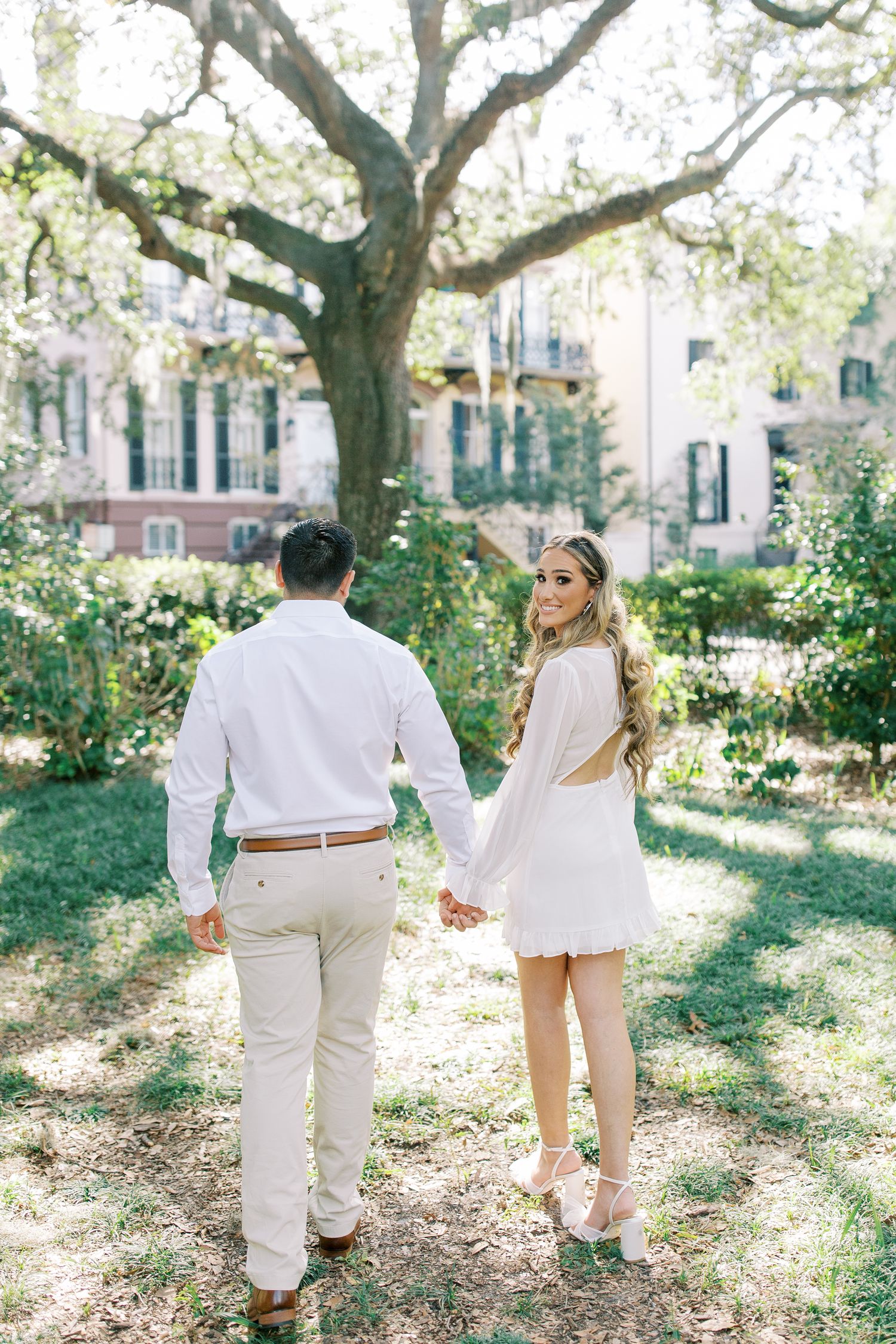 A romantic couple walks together in a sun-dappled garden courtyard with blooming trees and historic buildings in the background.