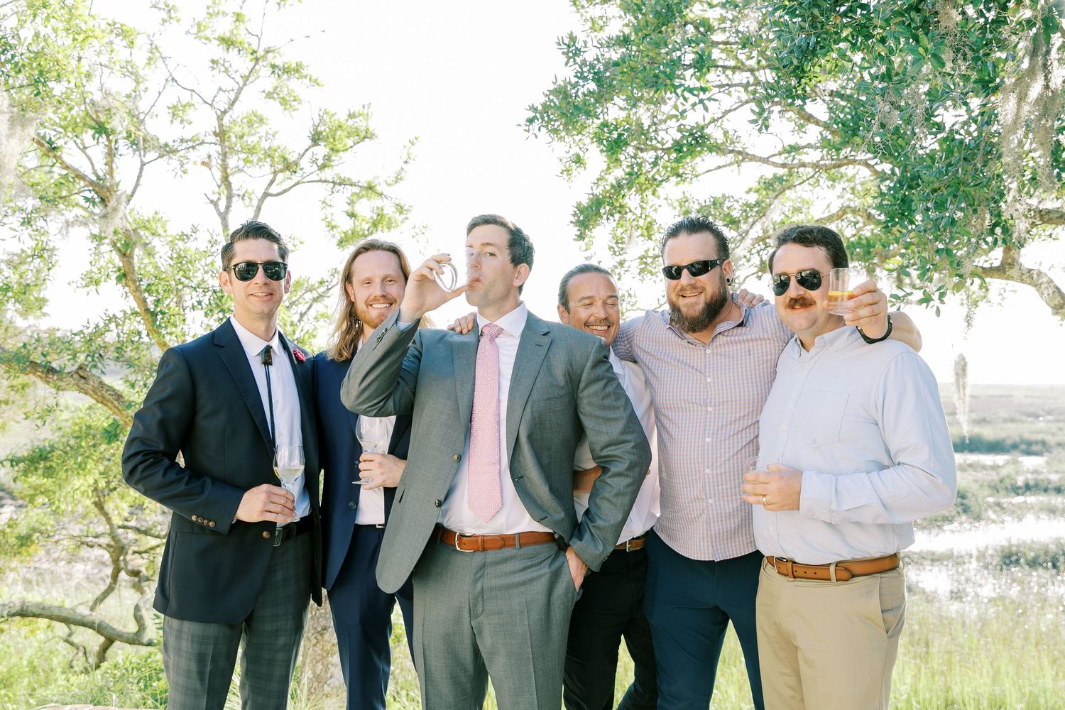 A group of wedding guests in formal attire pose together outdoors for a photo.