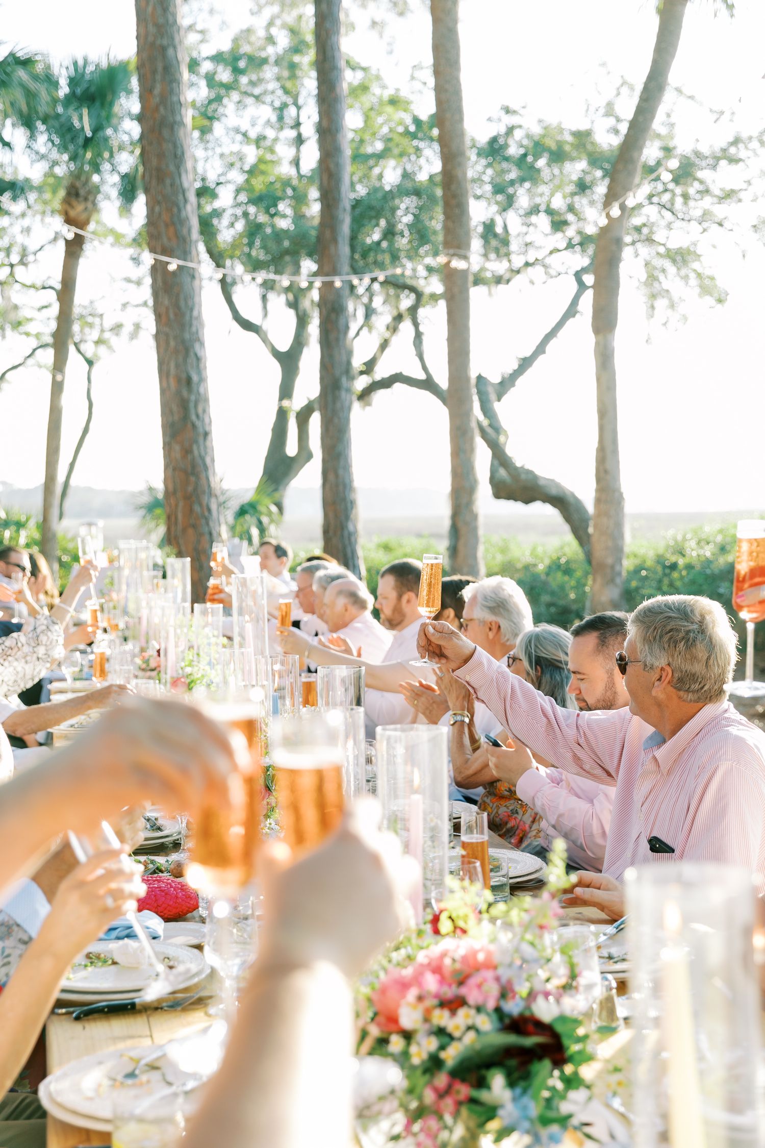 Wedding guests raise glasses in a toast at a long outdoor dinner table decorated with floral centerpieces.