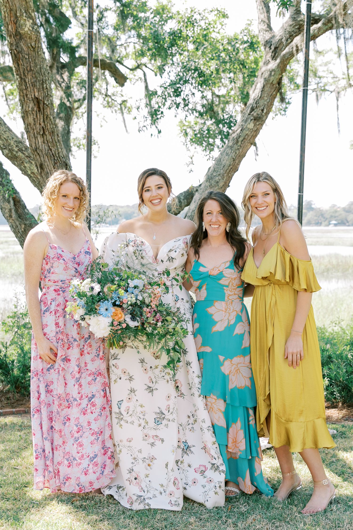 A group wearing floral dresses poses for a sunny outdoor photo under a large oak tree.