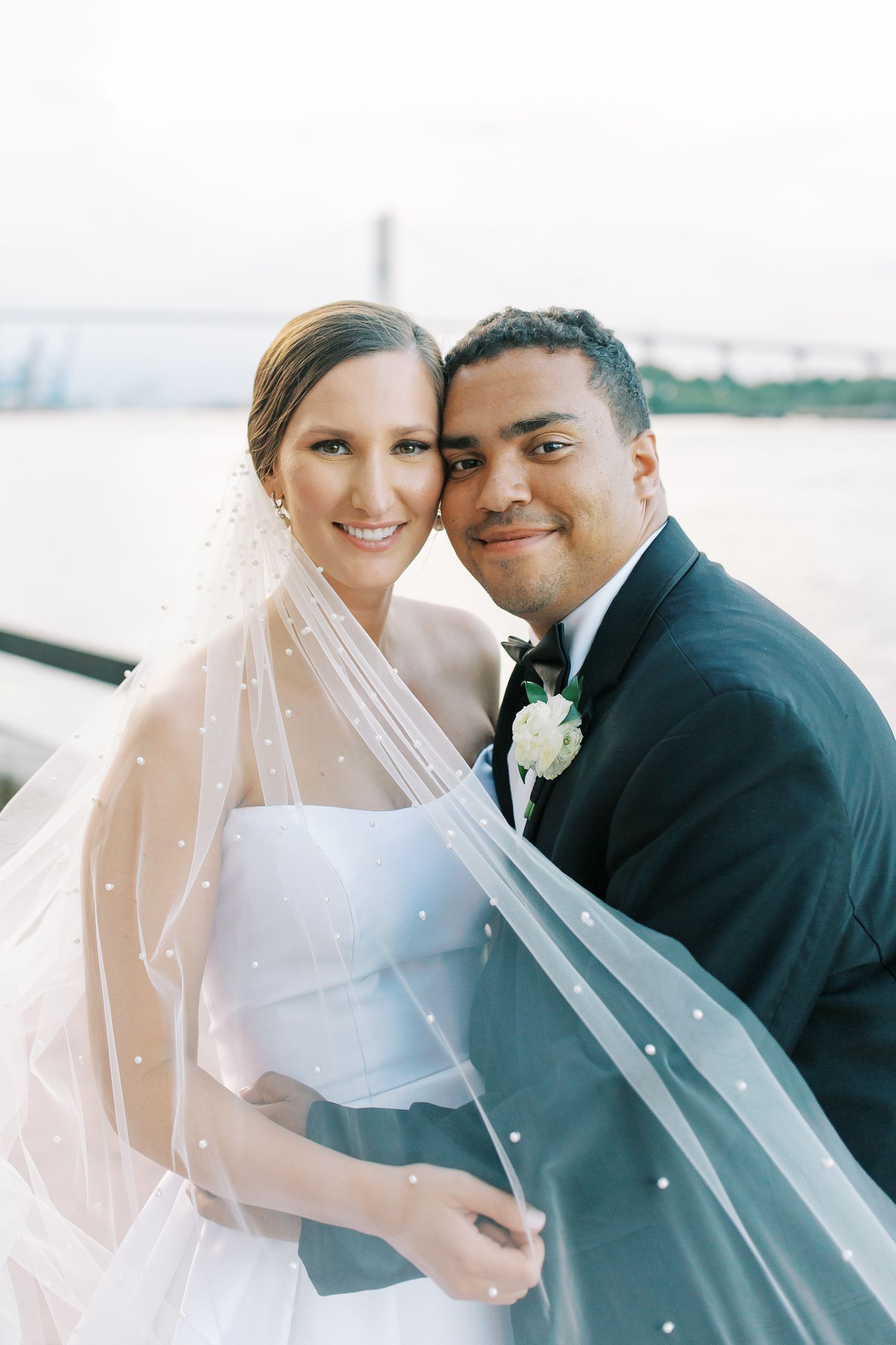 Wedding couple embraces outdoors with a waterfront backdrop and flowing veil.