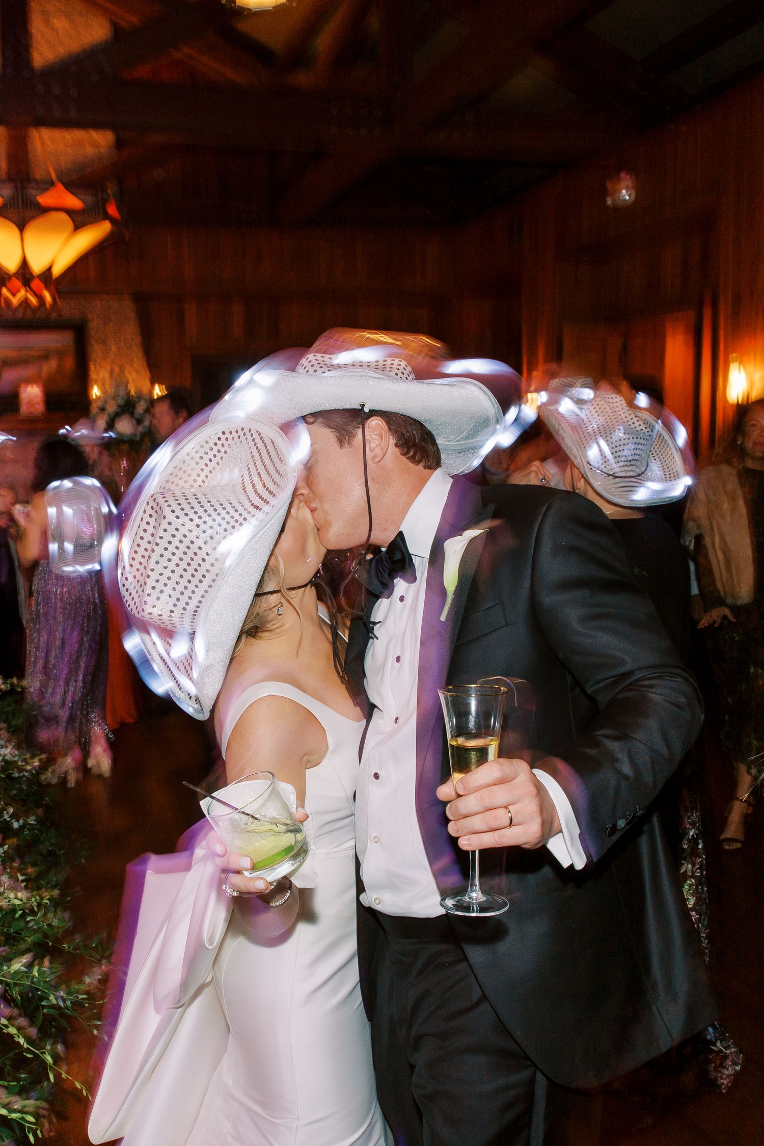 Wedding celebration with white cowboy hats and formal attire in a warmly lit wooden venue.