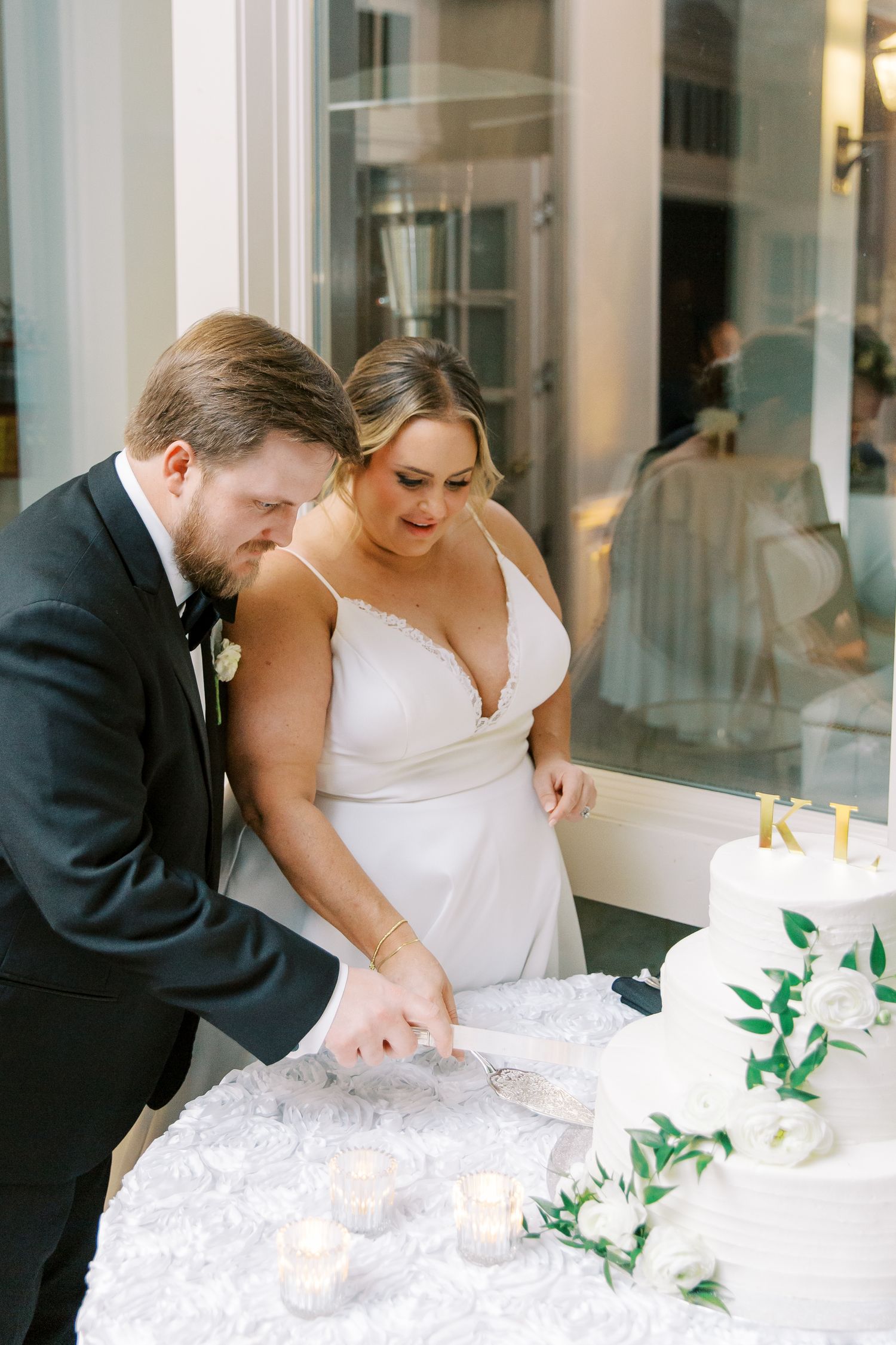 Newlyweds cut their white wedding cake decorated with greenery at their reception.
