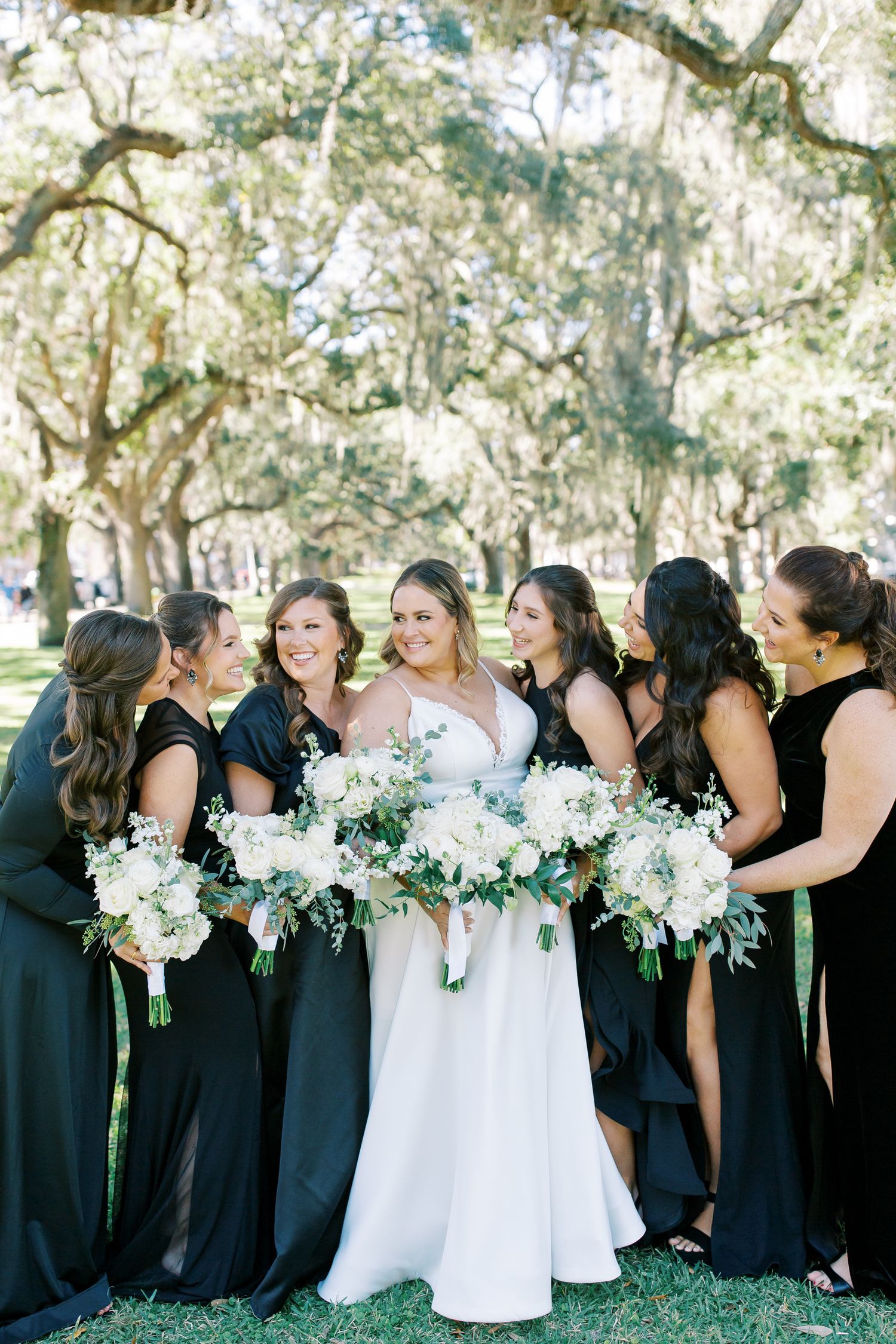A bridal party in dark gowns gathers outdoors under oak trees holding white bouquets.