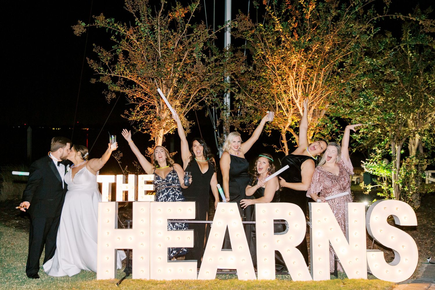 Wedding party poses with large illuminated 'THE HEARNS' letters at night with trees lit in background.