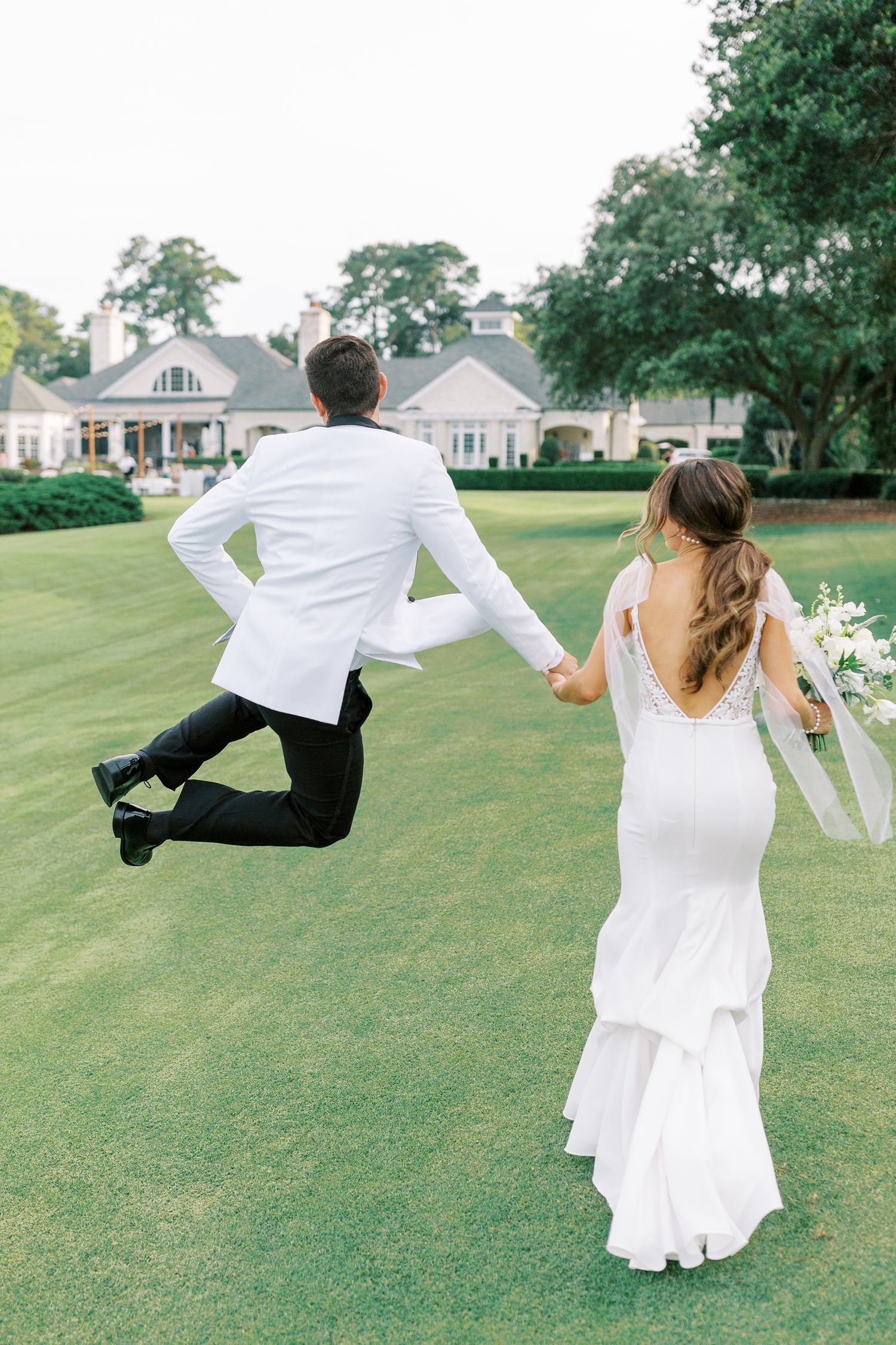 Playful wedding couple running and jumping together on a lush green lawn with houses in background.
