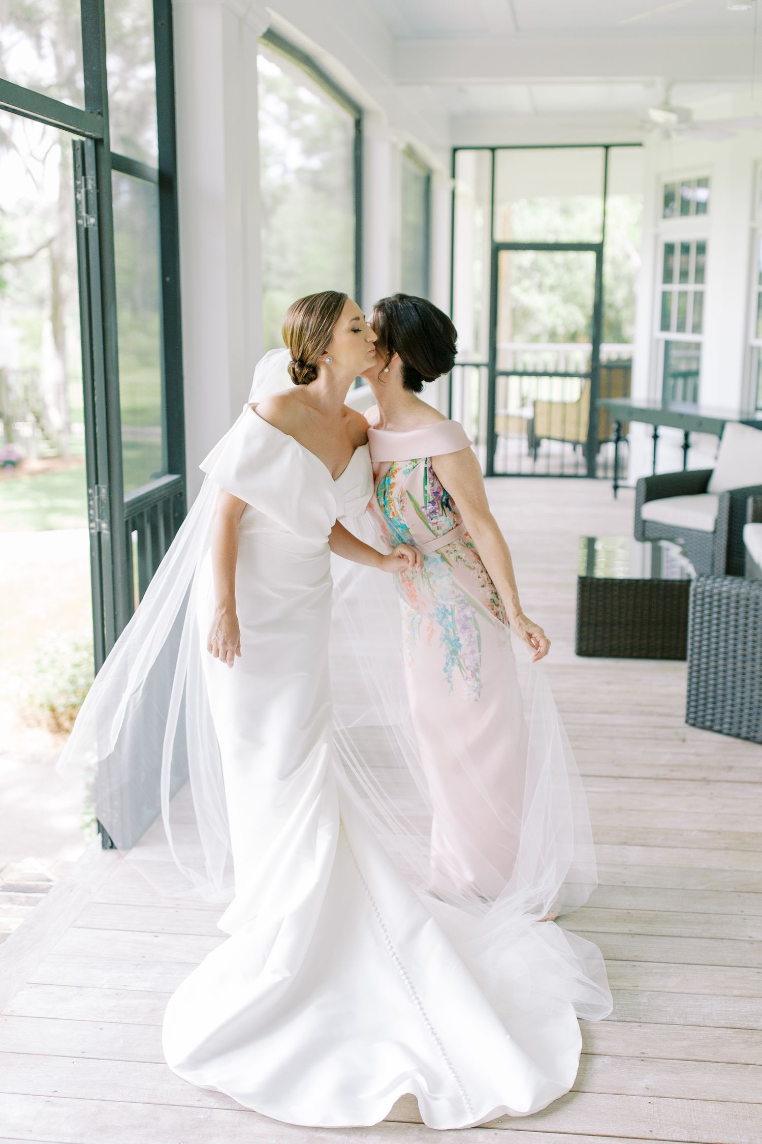 Two people in flowing white gowns share an intimate moment on a bright sunlit porch.