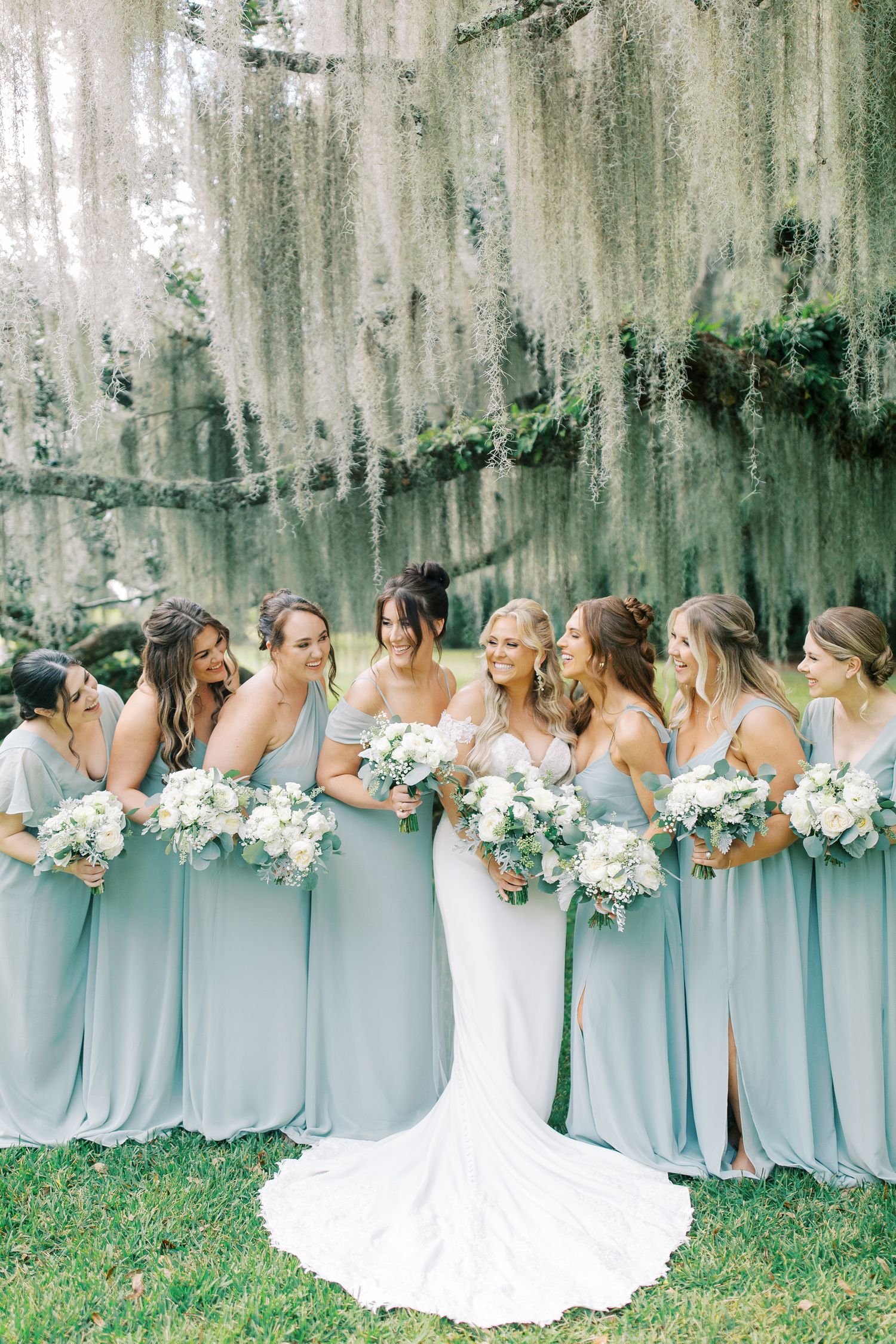 Bridal party in light blue dresses posing under hanging moss with white floral bouquets.