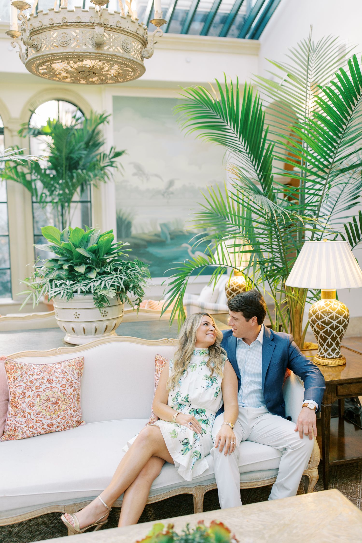 Elegant indoor sunroom decorated with palm plants, ornate chandelier and vintage furniture in pastel tones in the cloister at Sea Island.