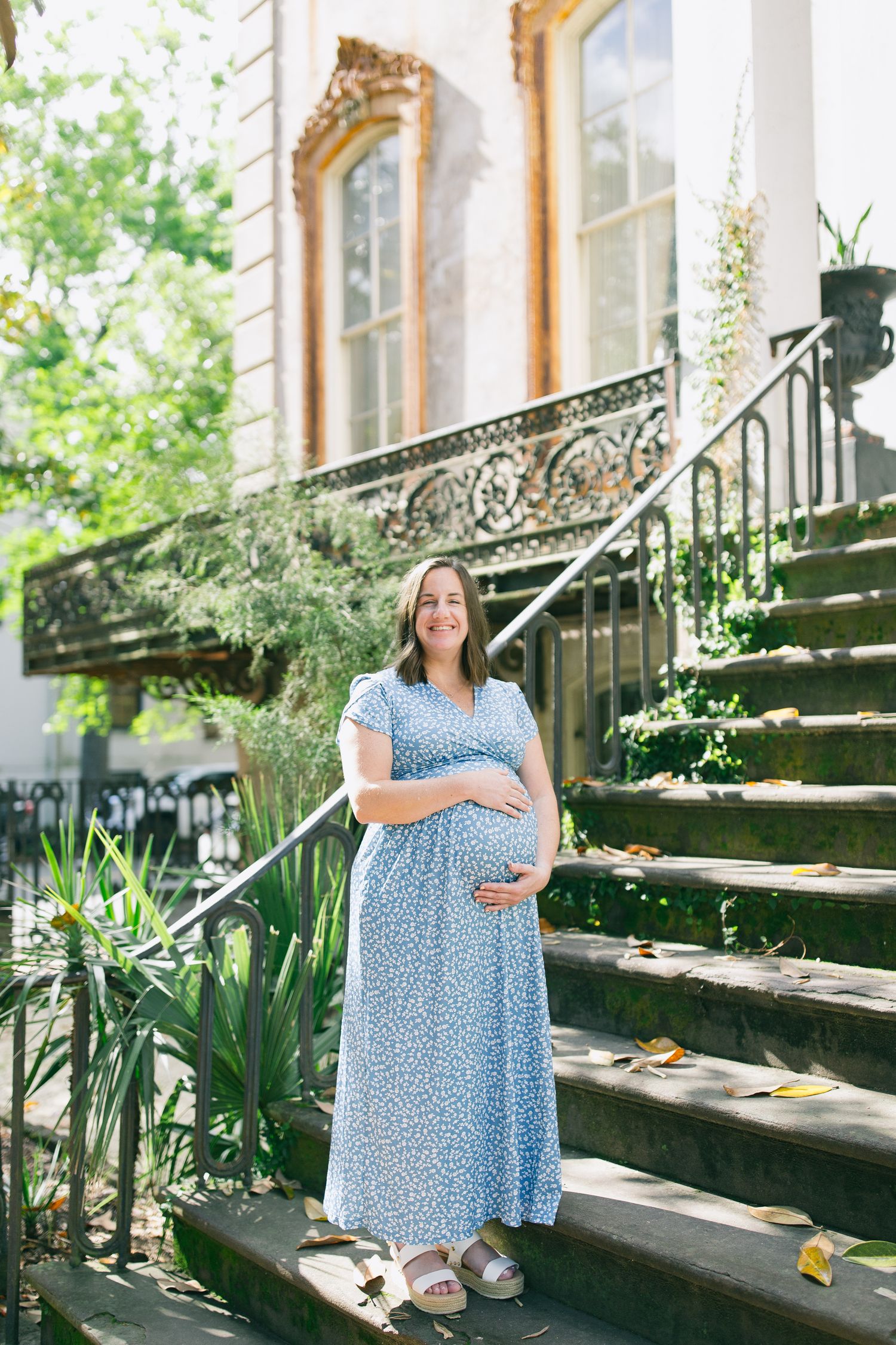 A woman in a blue floral dress stands on stone steps next to potted plants in a garden setting.