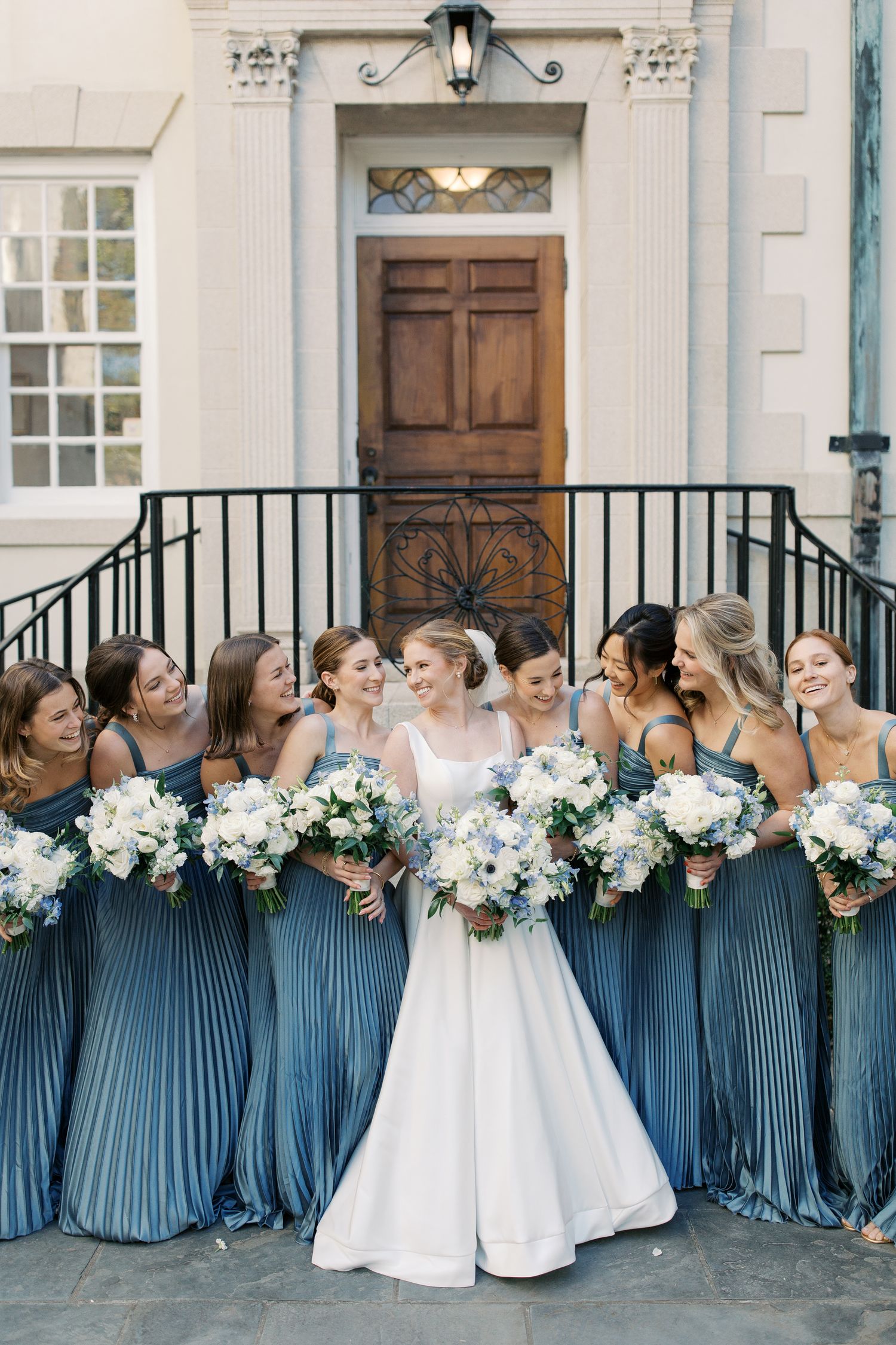 Large bridal party in blue dresses posing together outside traditional white church building.