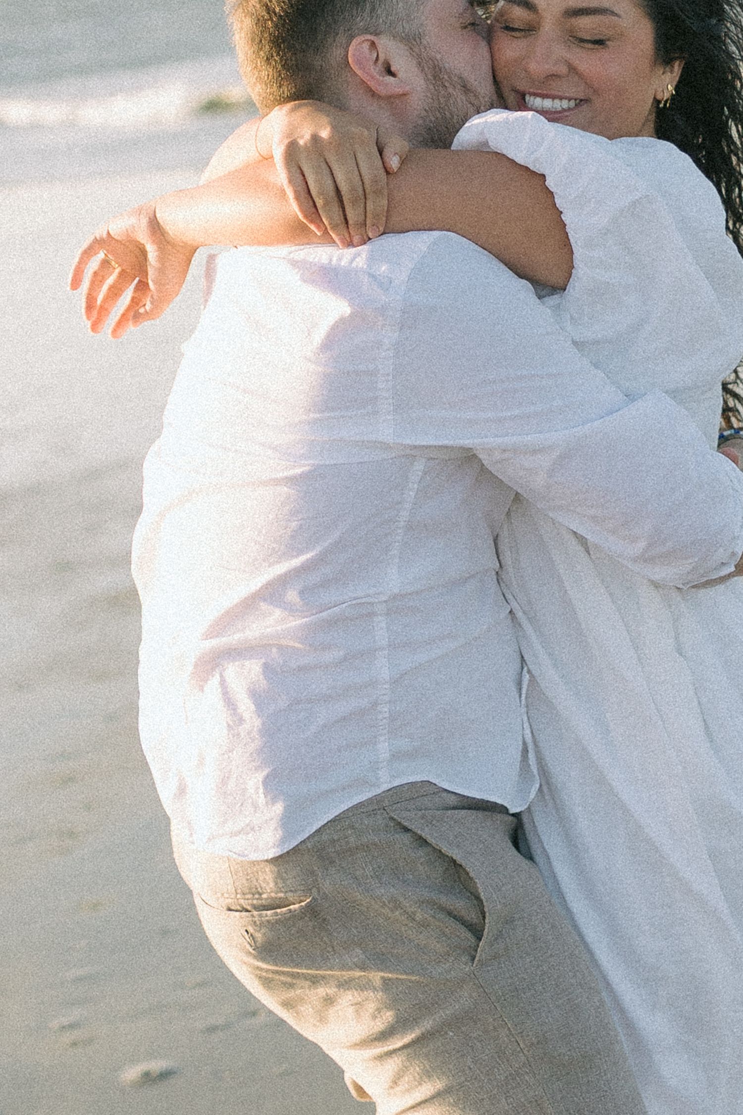 A romantic beach scene shows a couple in white embracing and dancing together on the sand at sunset.