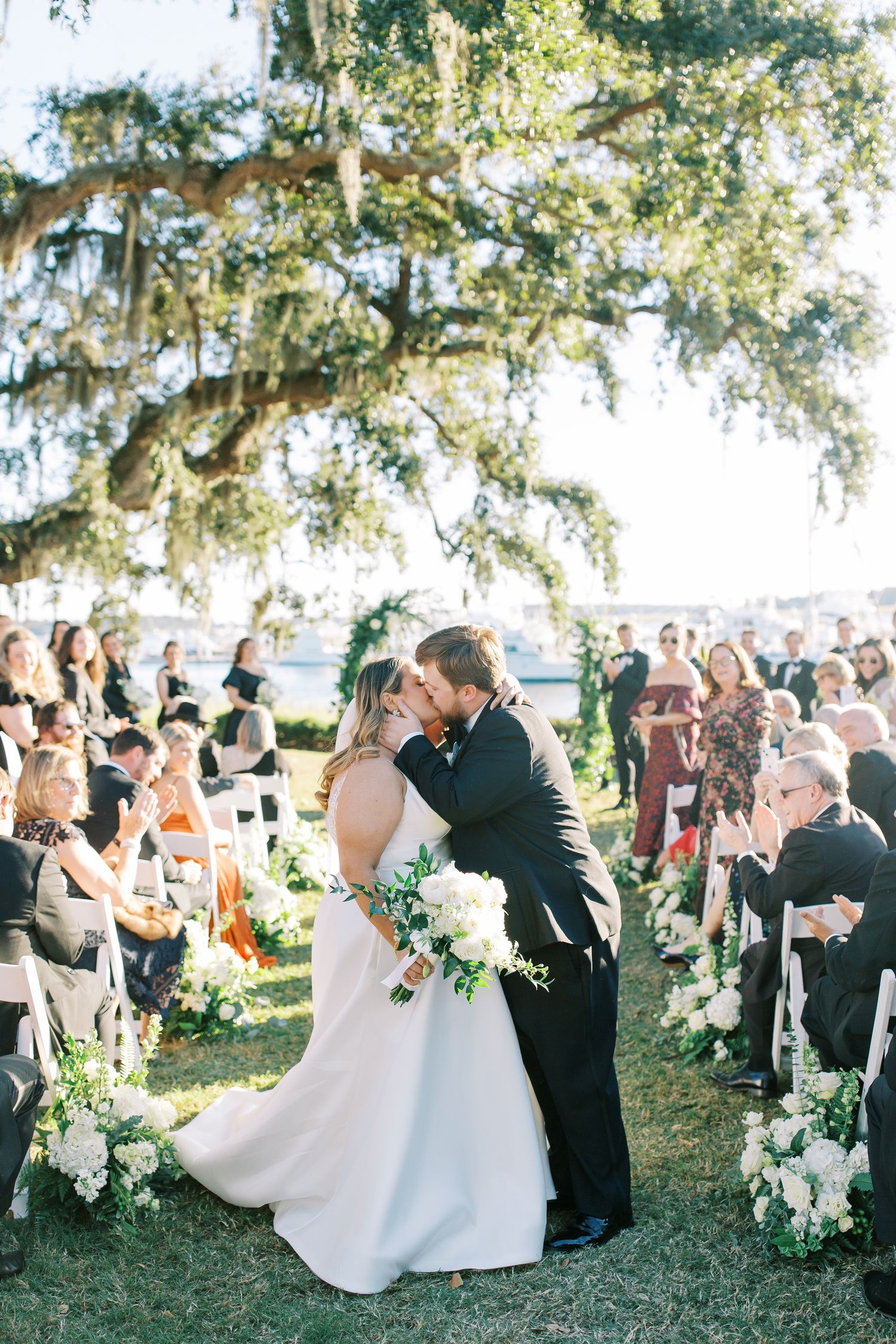 A romantic wedding ceremony takes place outdoors under a majestic oak tree draped with Spanish moss as guests look on.
