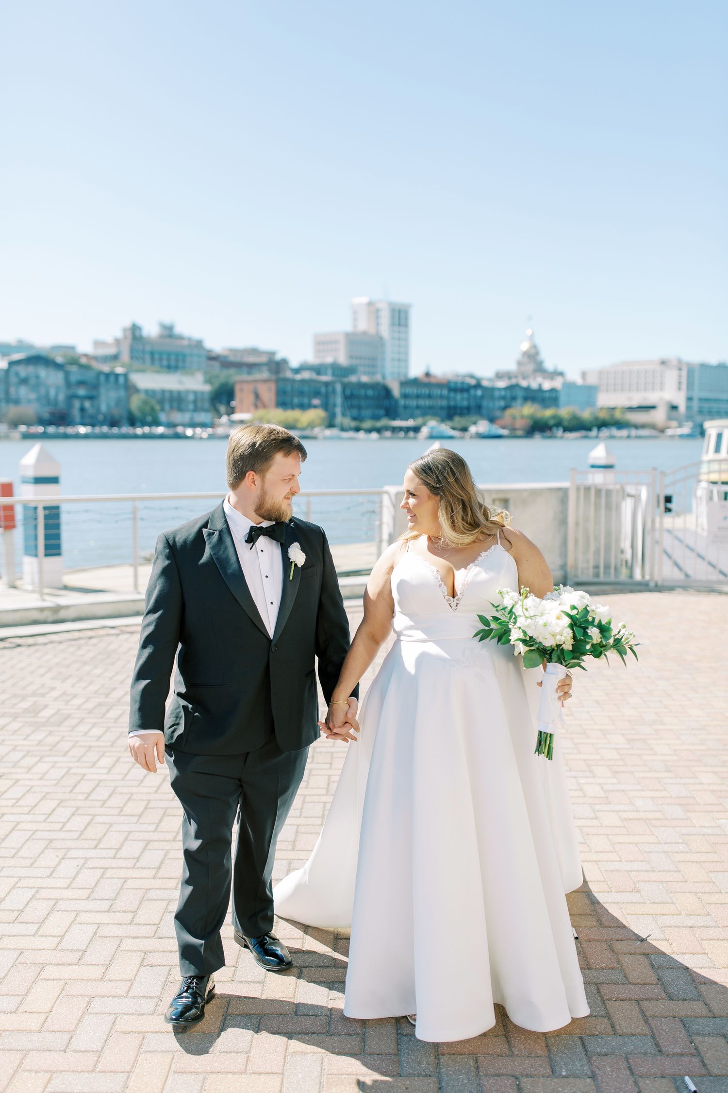 A bride in a white gown holds a bouquet while walking with her groom in a tuxedo along a waterfront promenade.