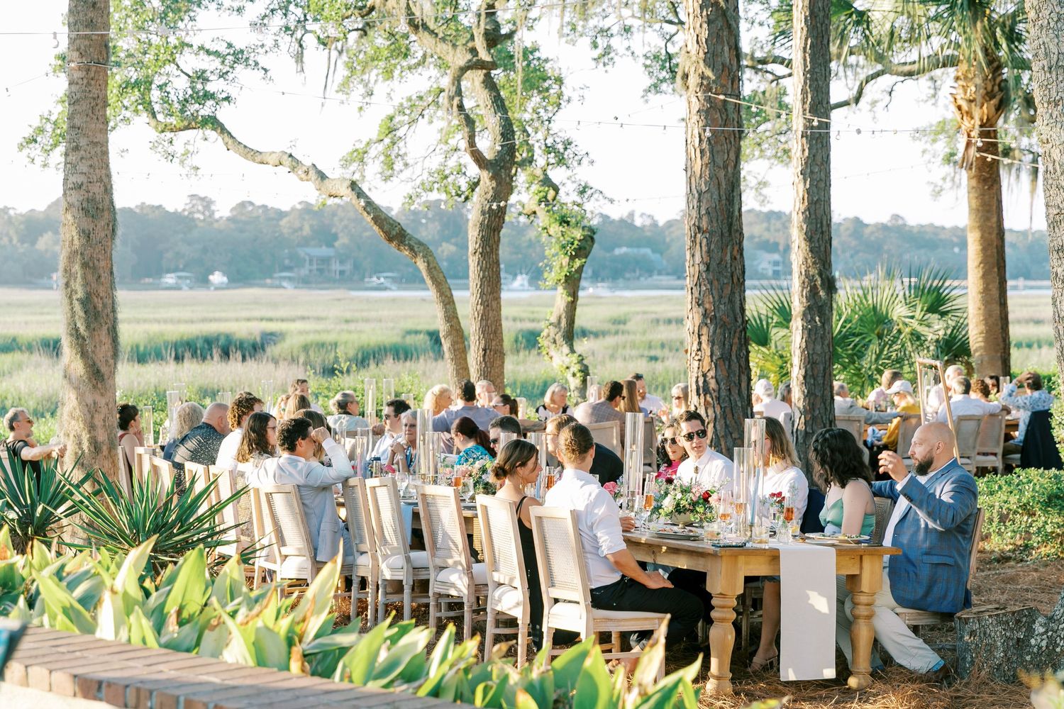 Outdoor wedding reception dining setup under oak trees with string lights.