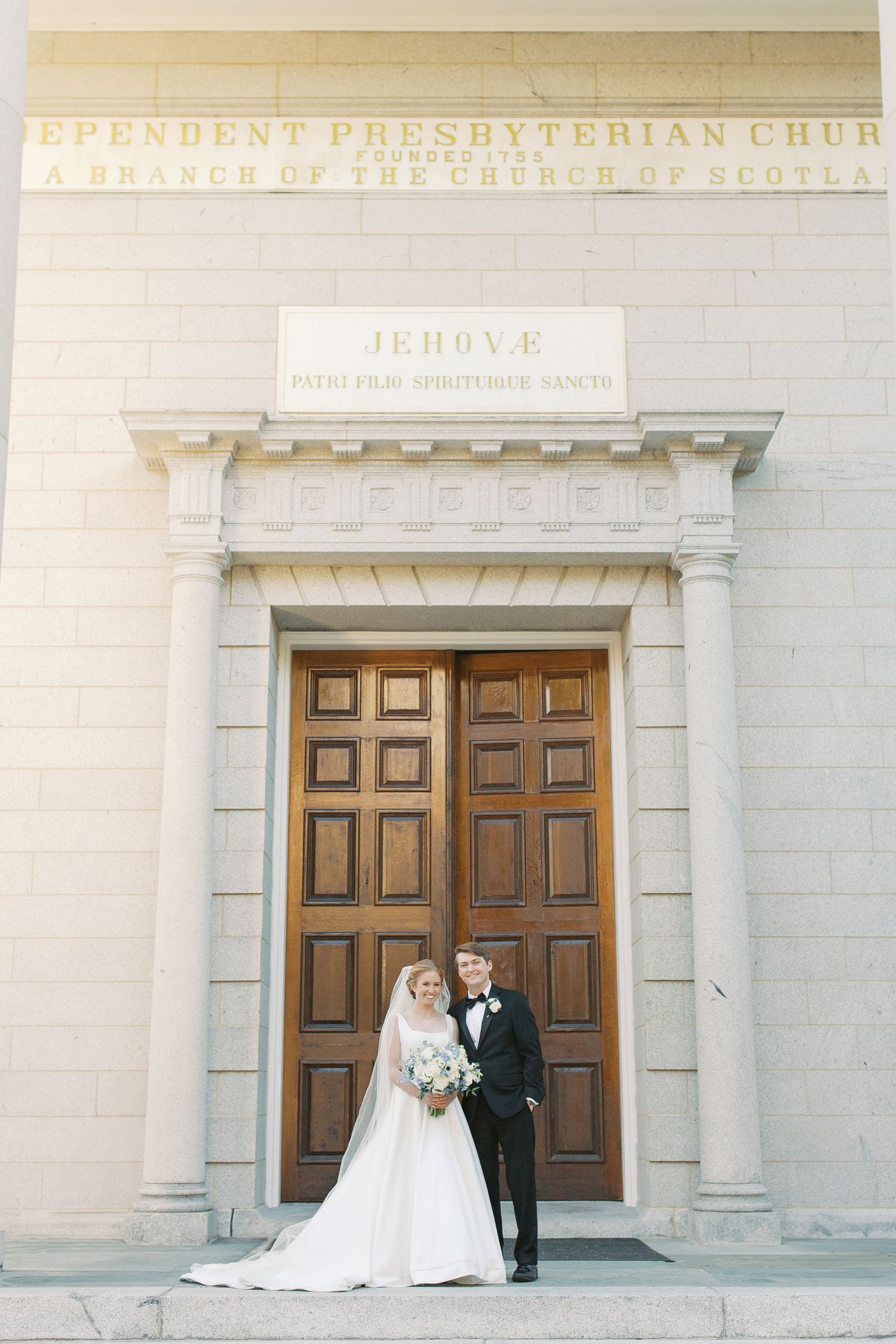 Classic white church facade with wooden doors serves as backdrop for formal wedding portraits.