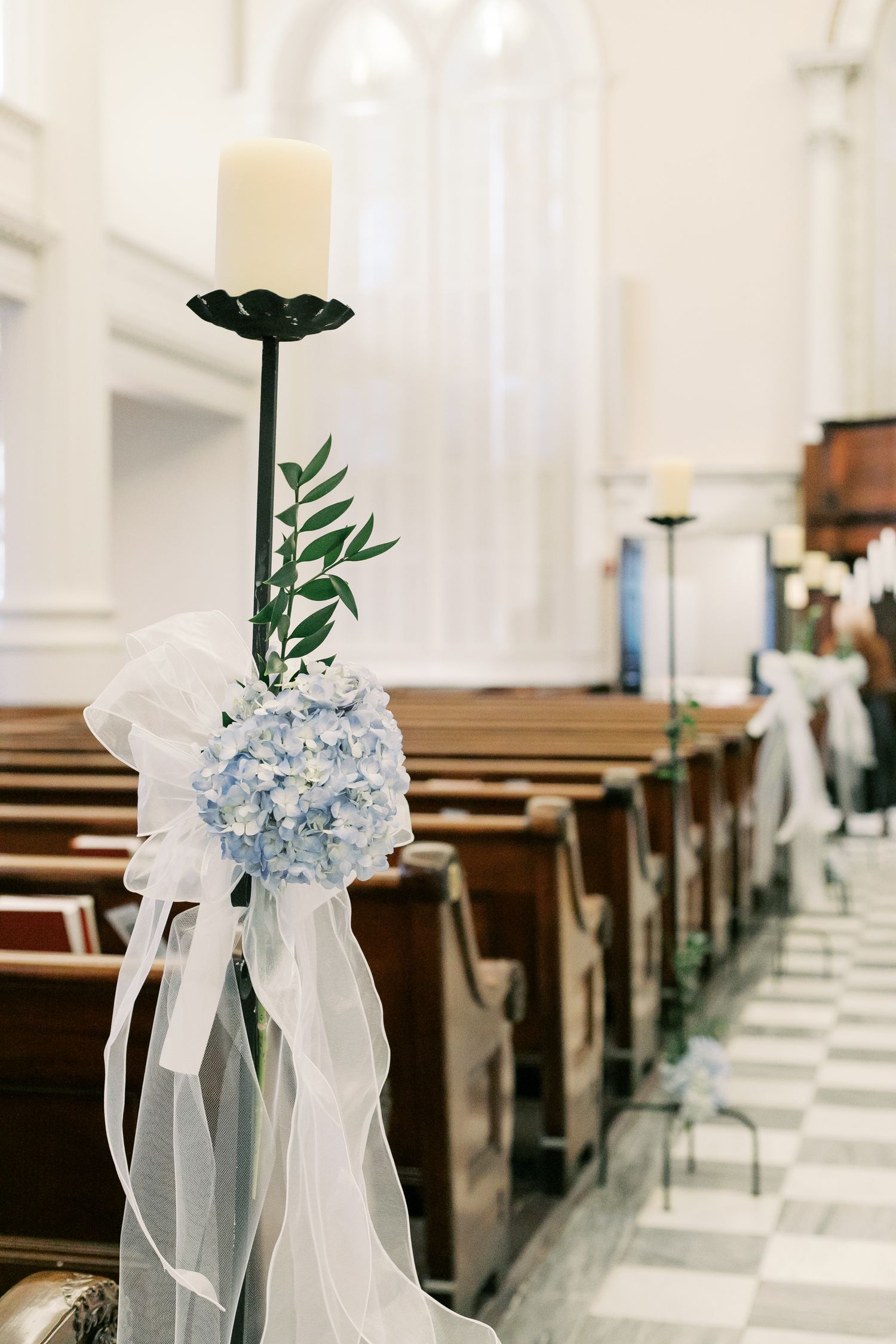 Church pew decorated with white candle, blue hydrangea, and sheer white ribbon for wedding ceremony.