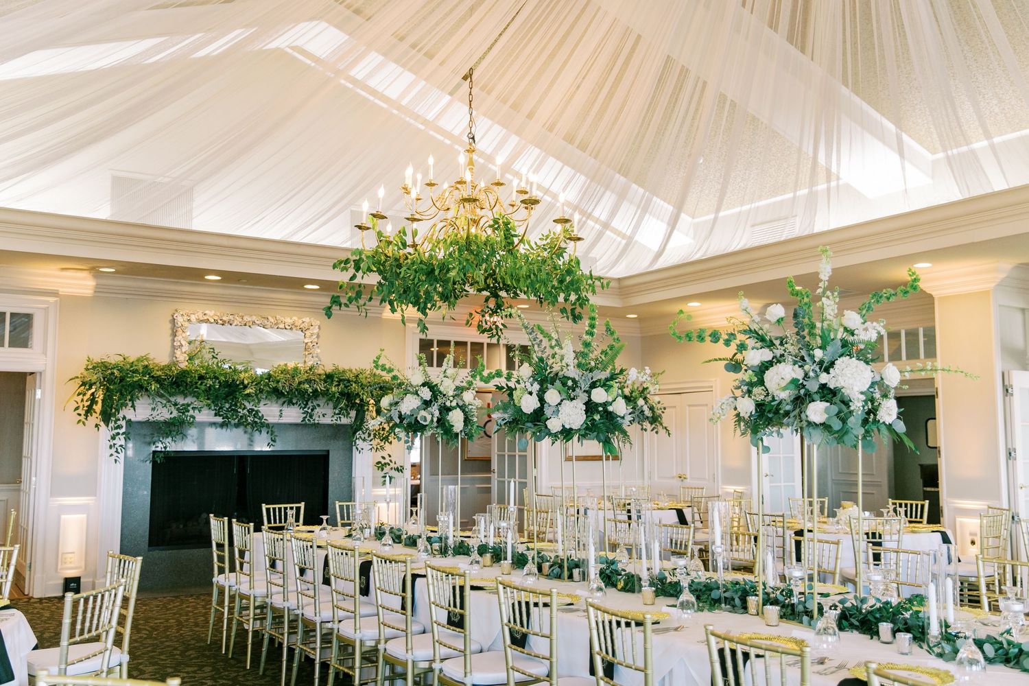Elegant indoor wedding reception venue with draped ceiling and lush greenery centerpieces on white tables.