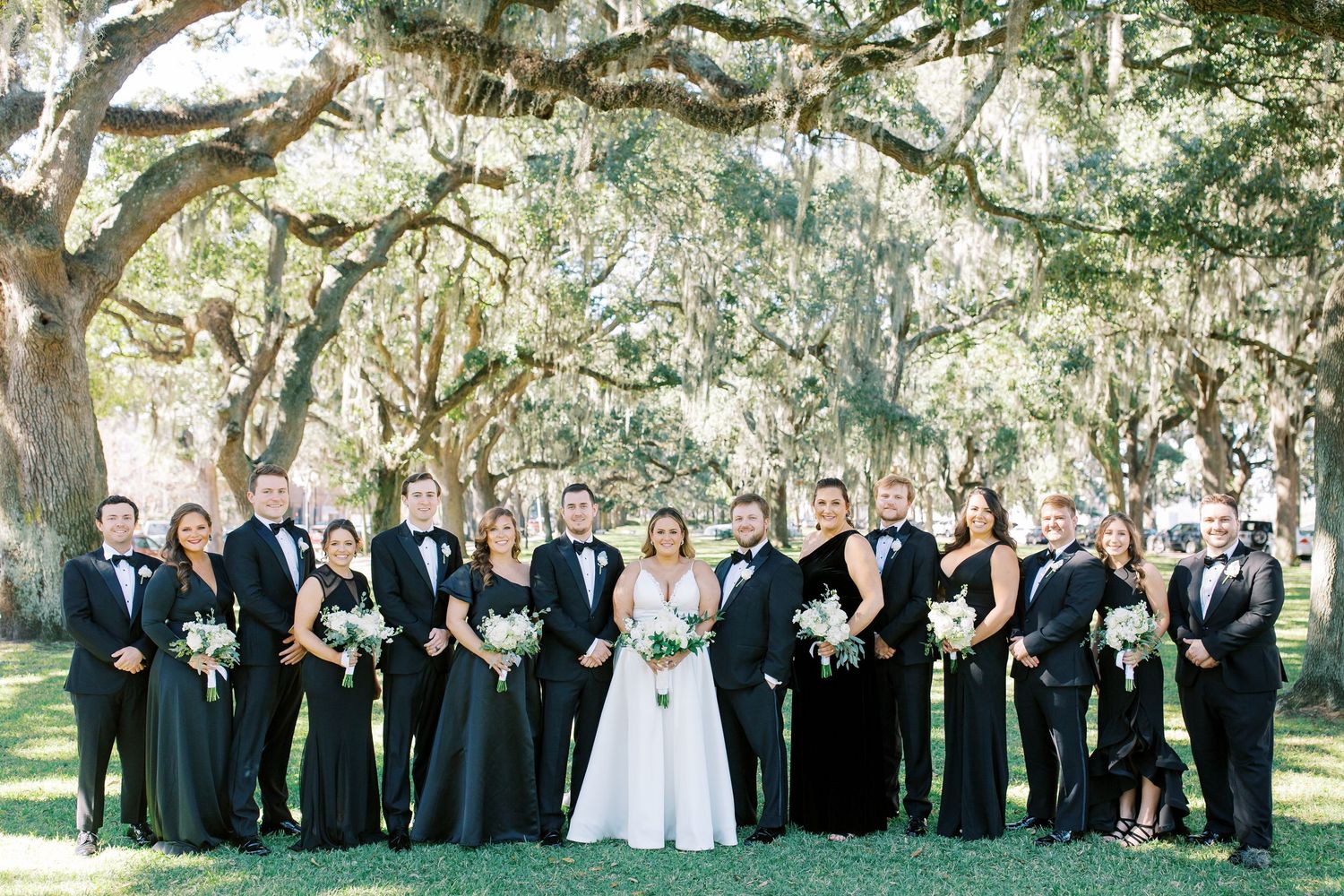 Wedding party in formal attire poses under Spanish moss-draped trees on a sunny day.