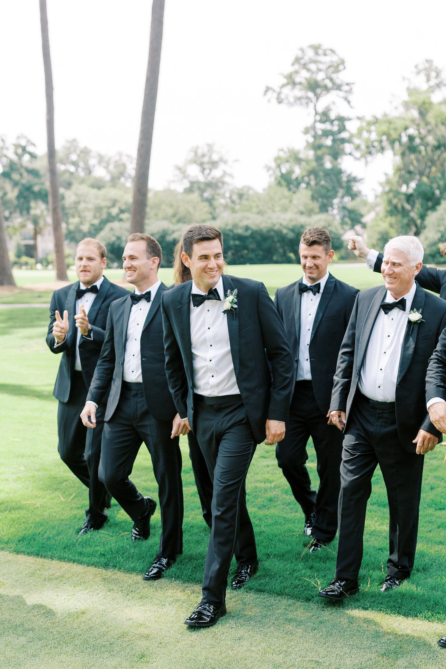 Groomsmen in black tuxedos walking together on a manicured lawn.