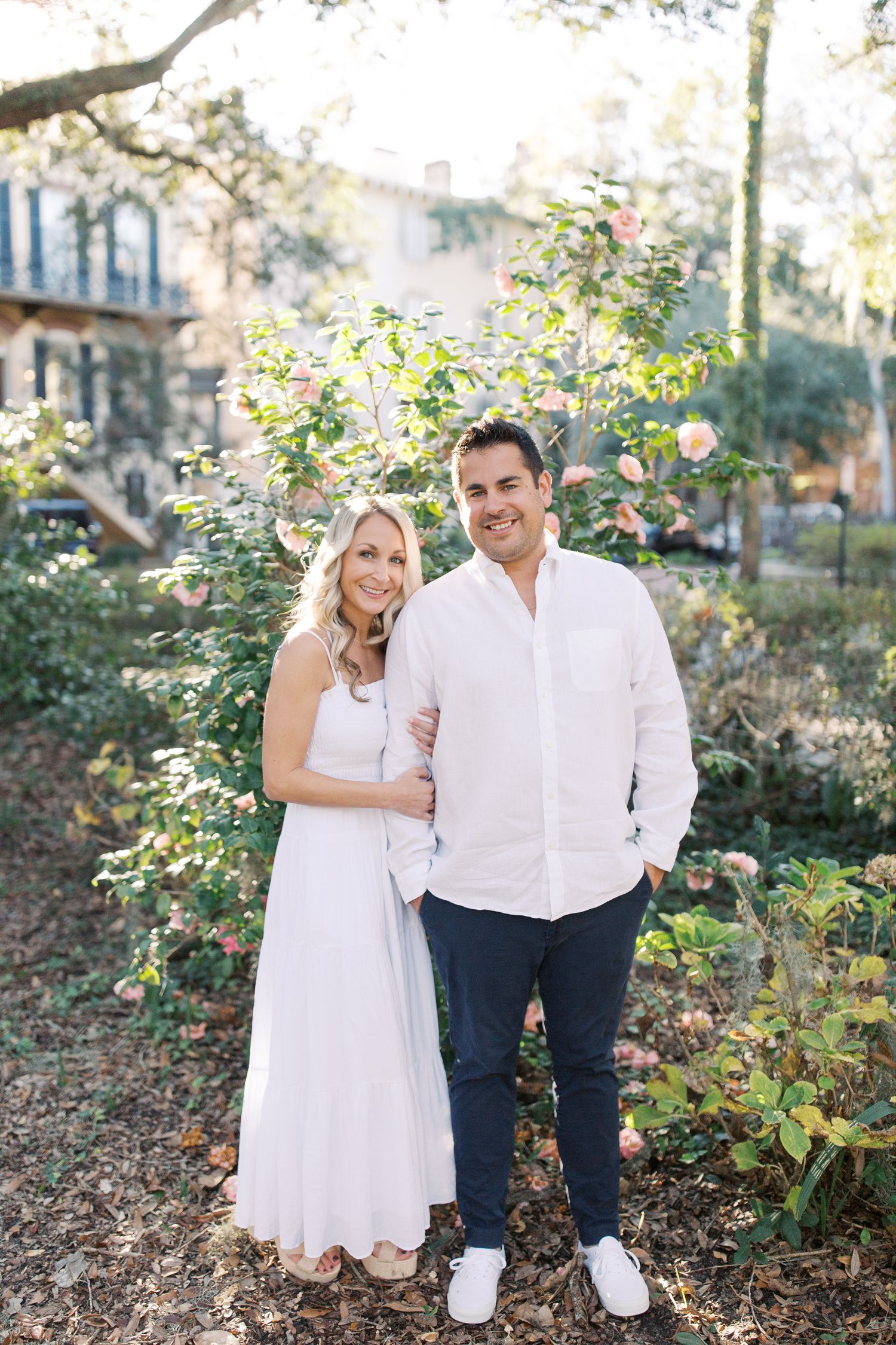 Engagement portrait of couple in white attire standing in lush garden setting.