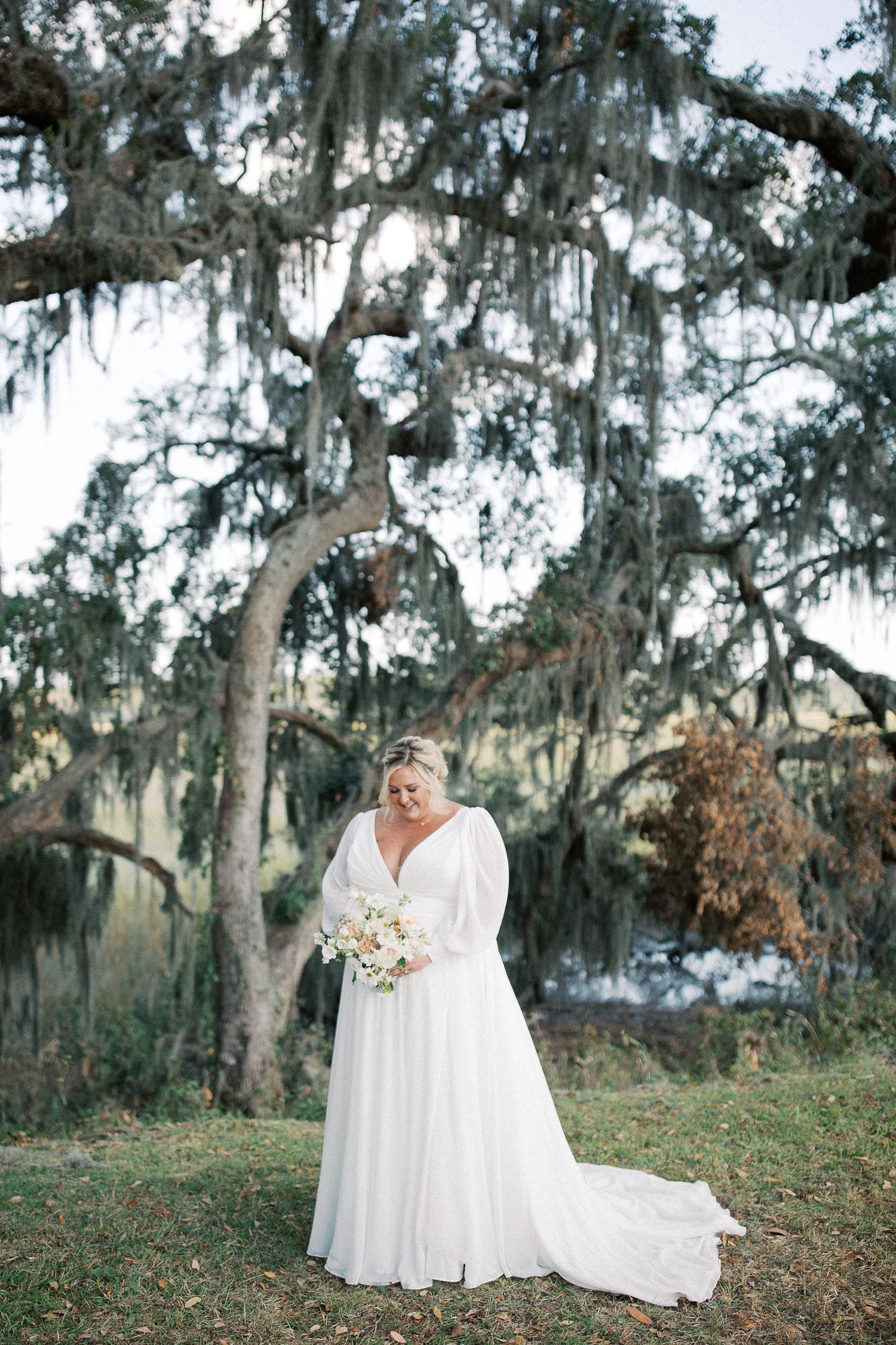 Elegant bride in flowing white gown poses beneath Spanish moss covered trees at Bethesda Academy.