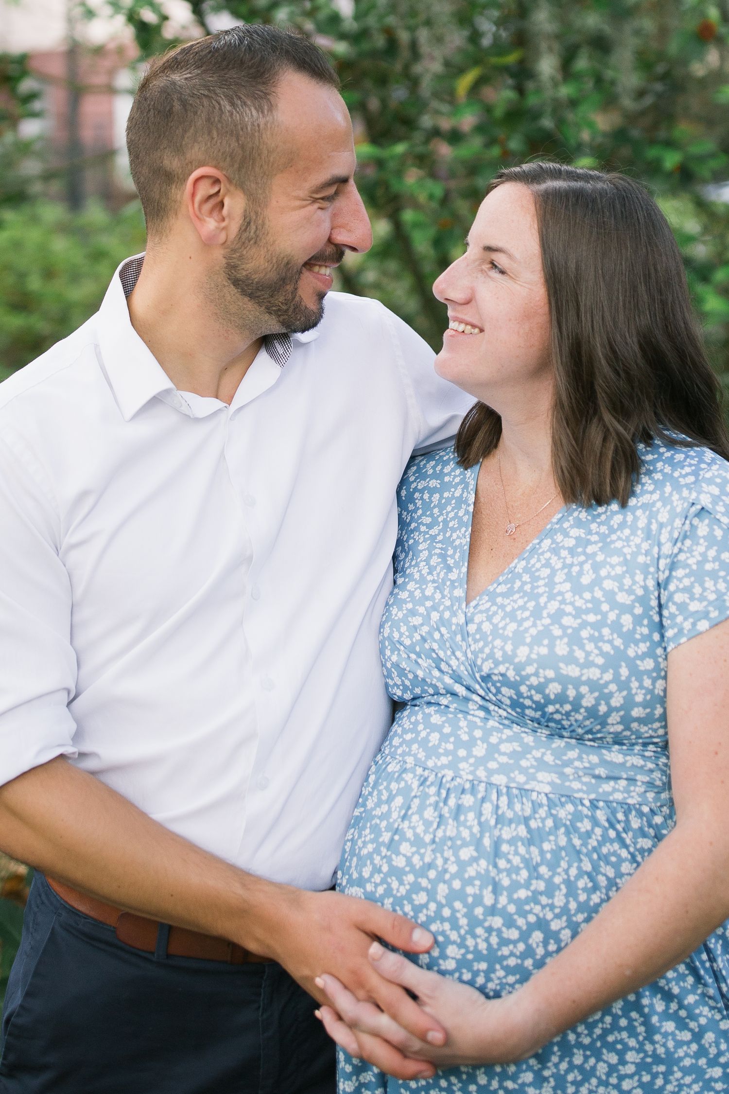 Expectant couple embraces lovingly outdoors wearing white and blue floral dress in a romantic maternity photoshoot.
