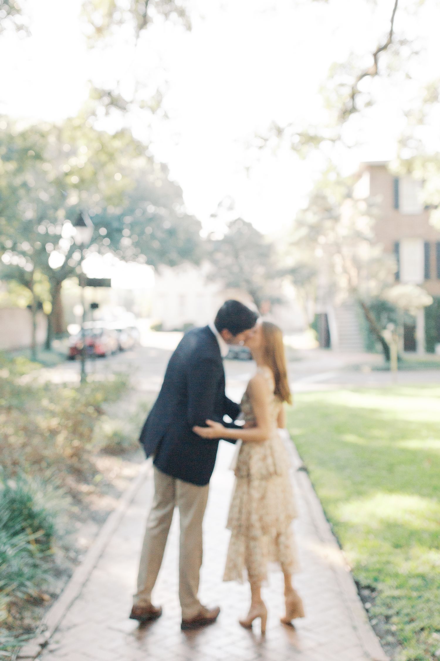 A romantic couple share a tender moment while walking on a sidewalk path beneath trees on a sunny day.