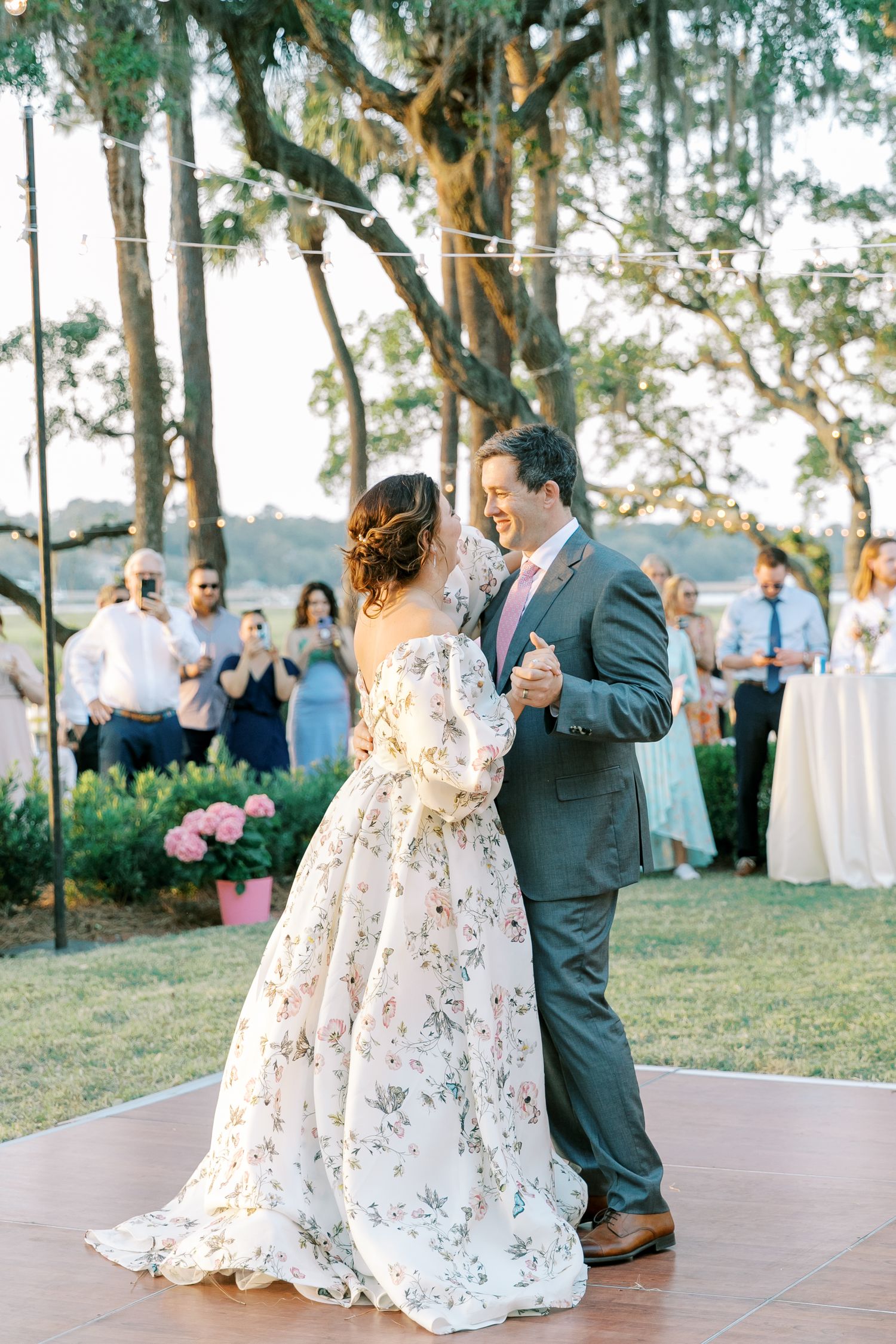A couple shares their first dance on an outdoor dance floor surrounded by palm trees and wedding guests.