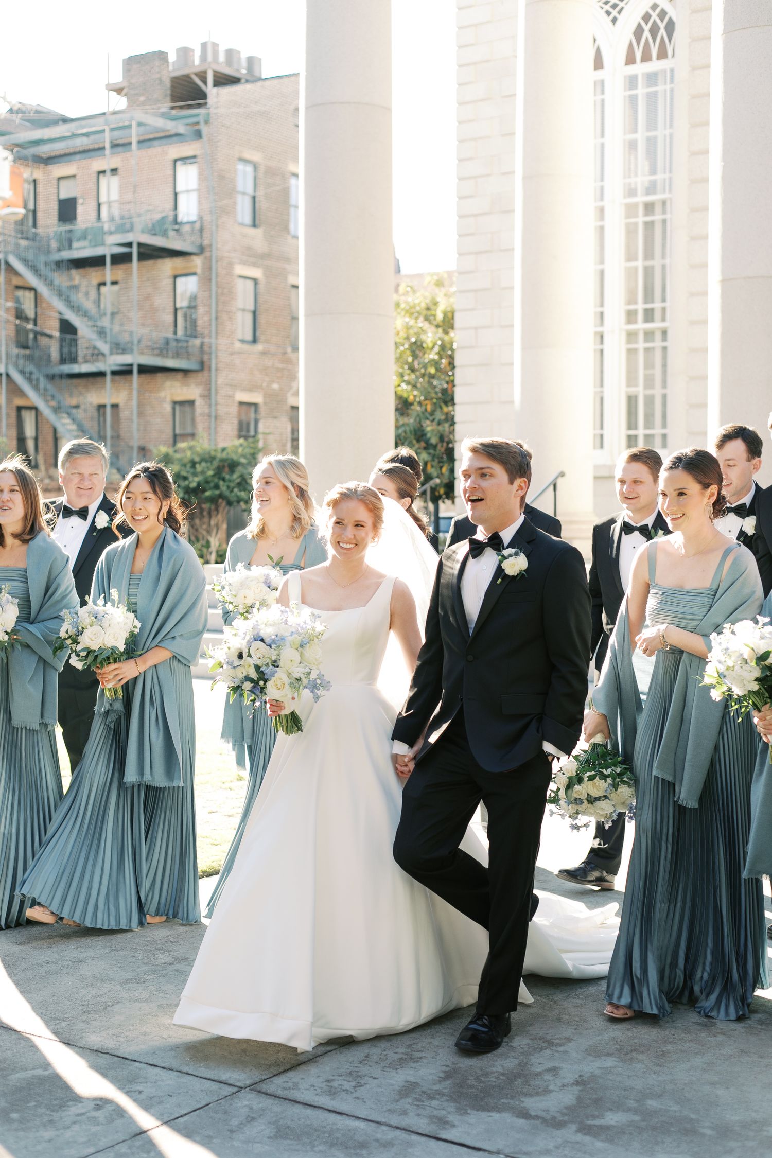 Wedding party in dusty blue bridesmaid dresses and black tuxedos posing by white columns.