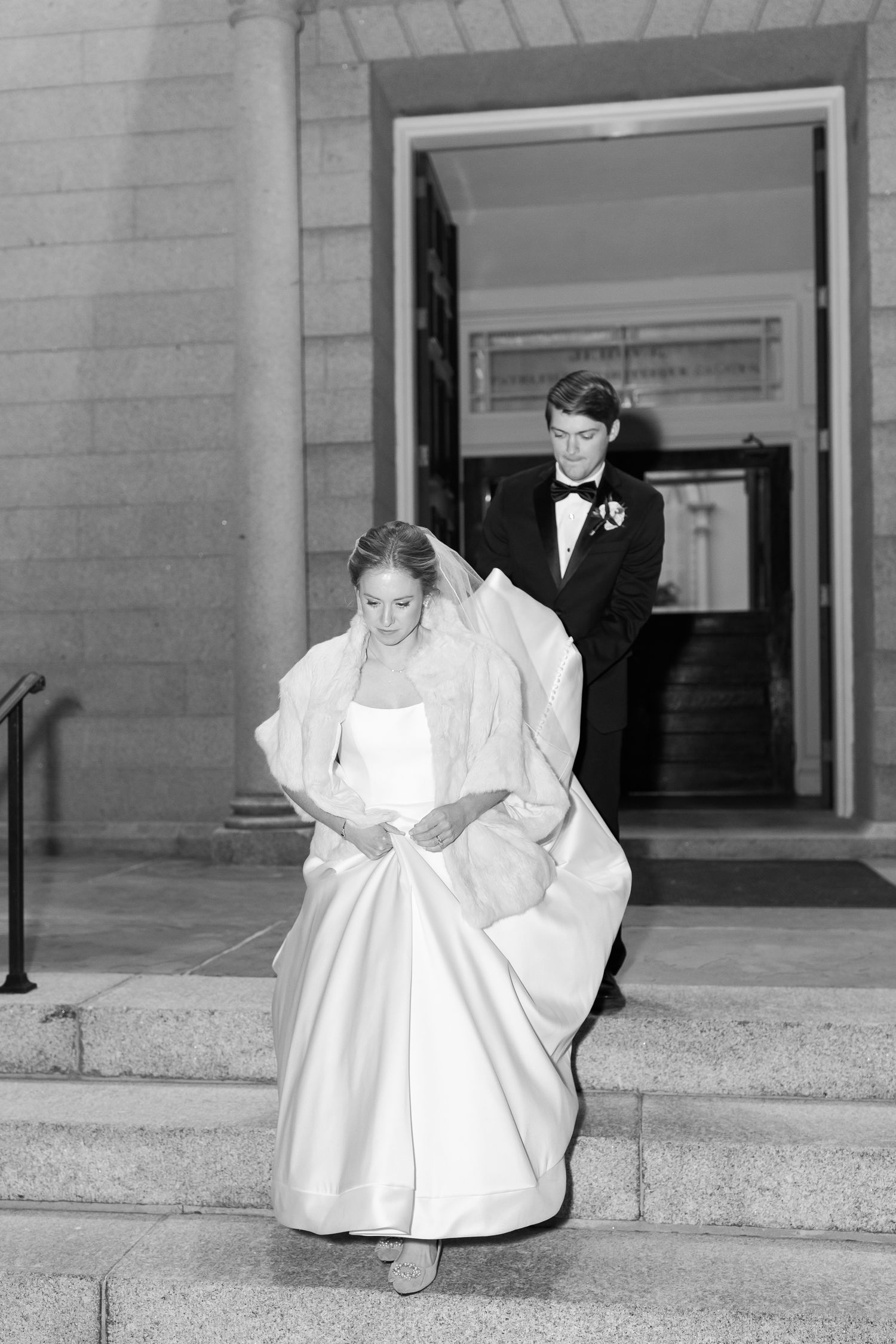 Black and white photo of wedding couple descending stone steps in formal attire.