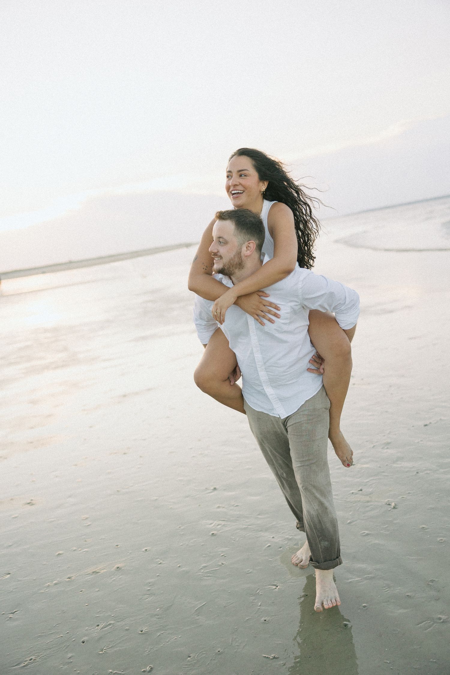A couple plays and laughs together on a beach at sunset giving piggyback rides.
