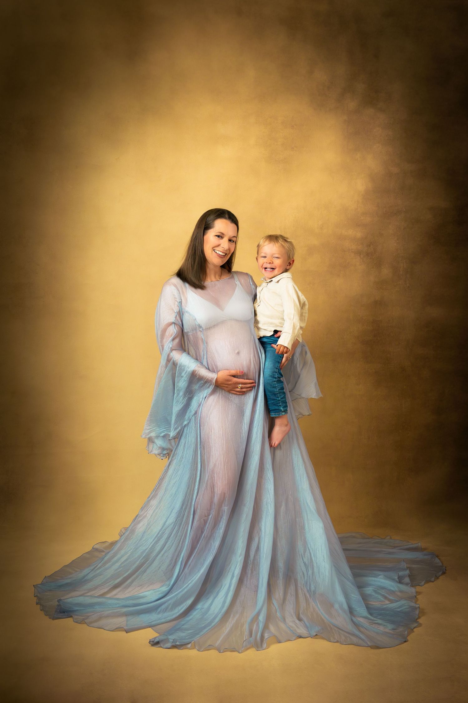 Gravidfotograf i studio Göteborg – mamma i vacker blå klänning med barn i famnen framför handmålad bakgrund.