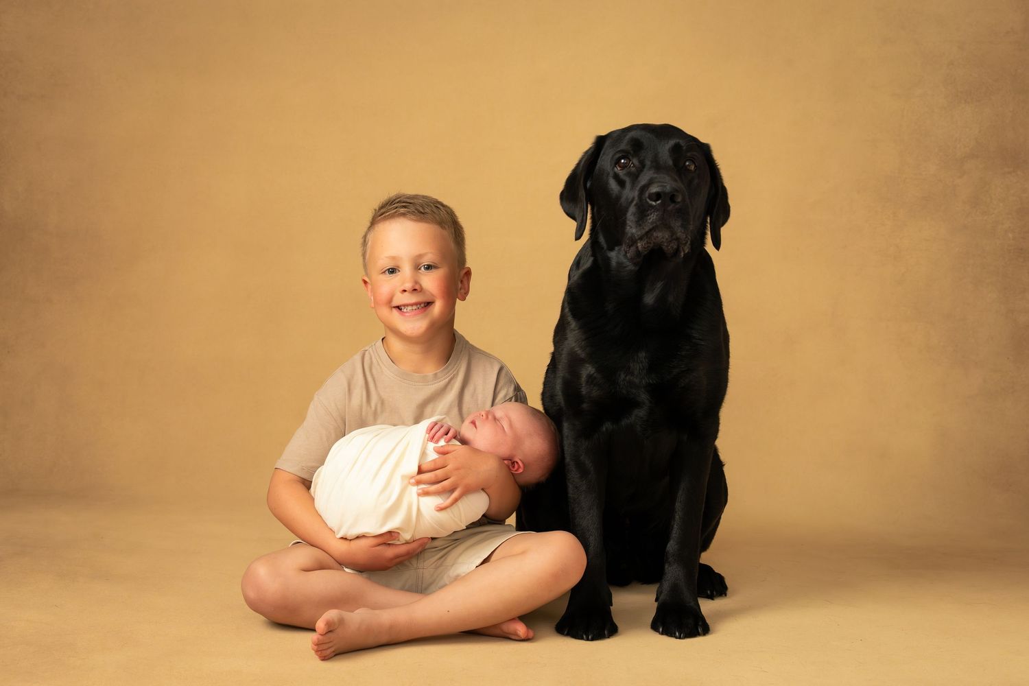 Syskon håller om sin nyfödda lillasyster och deras hund sitter bredvid under familjefotografering i studio.