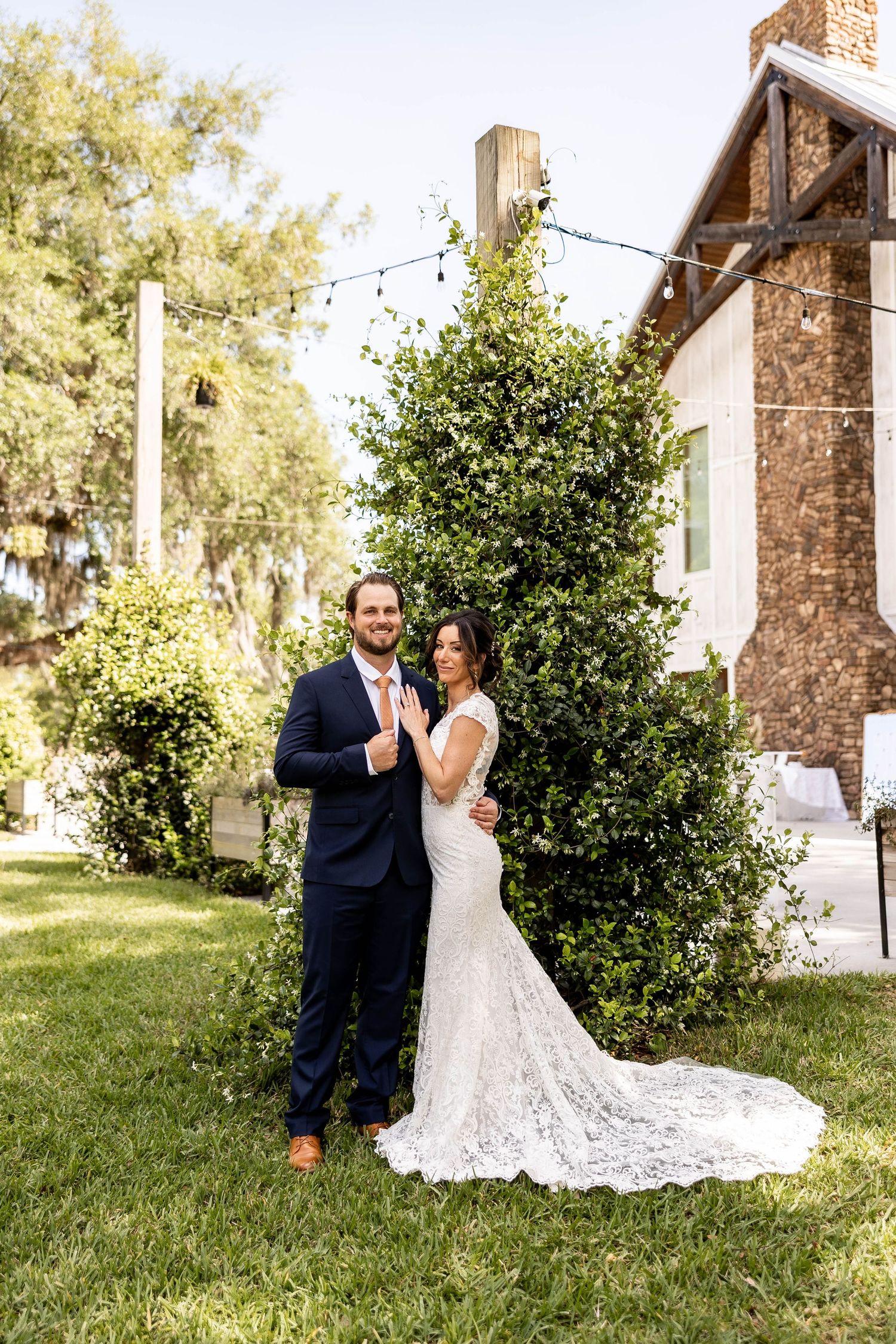 Bride in lace gown and groom in navy suit pose together on green lawn beside lush shrub at outdoor wedding venue.