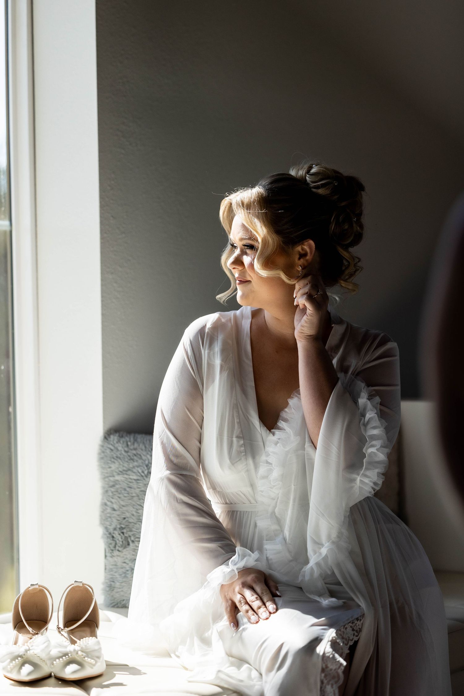 A bride in a white robe sits by a window in soft natural light with wedding shoes nearby.