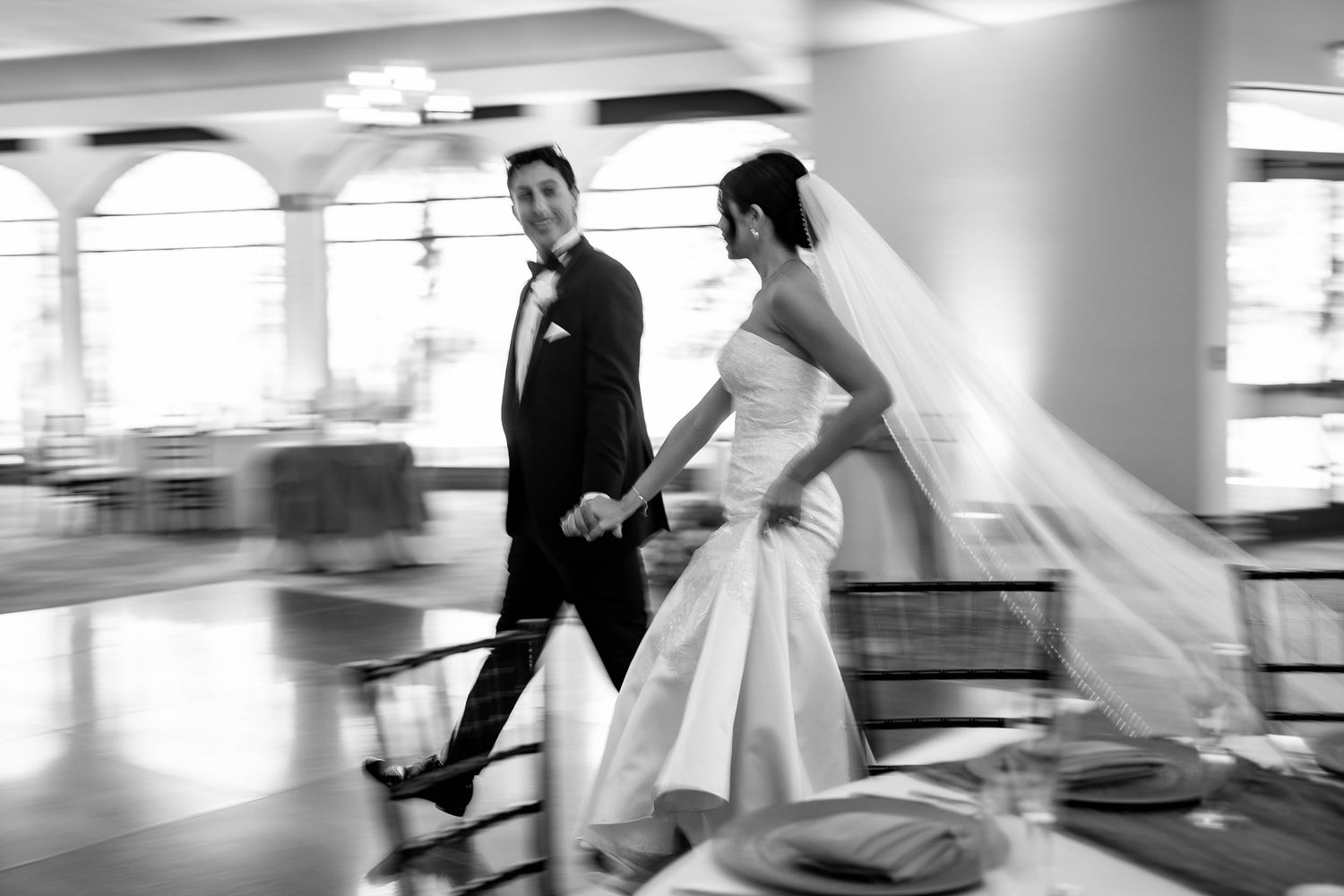 Black and white photo of a bride and groom holding hands walking into their wedding reception venue.