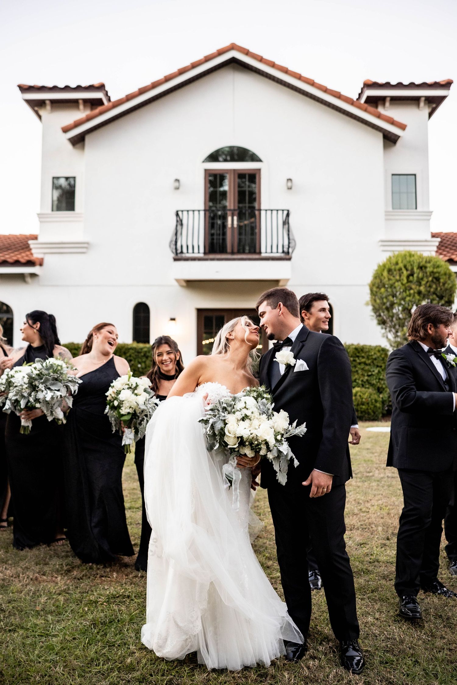 Bride and groom share a romantic moment outdoors, surrounded by wedding party in black attire, with white bouquets near elegant venue.