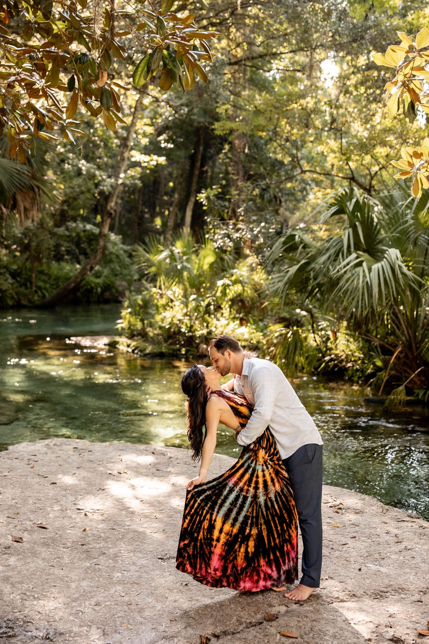A romantic moment shared by a couple in a colorful dress and white shirt by a serene forest stream on a sunny day.