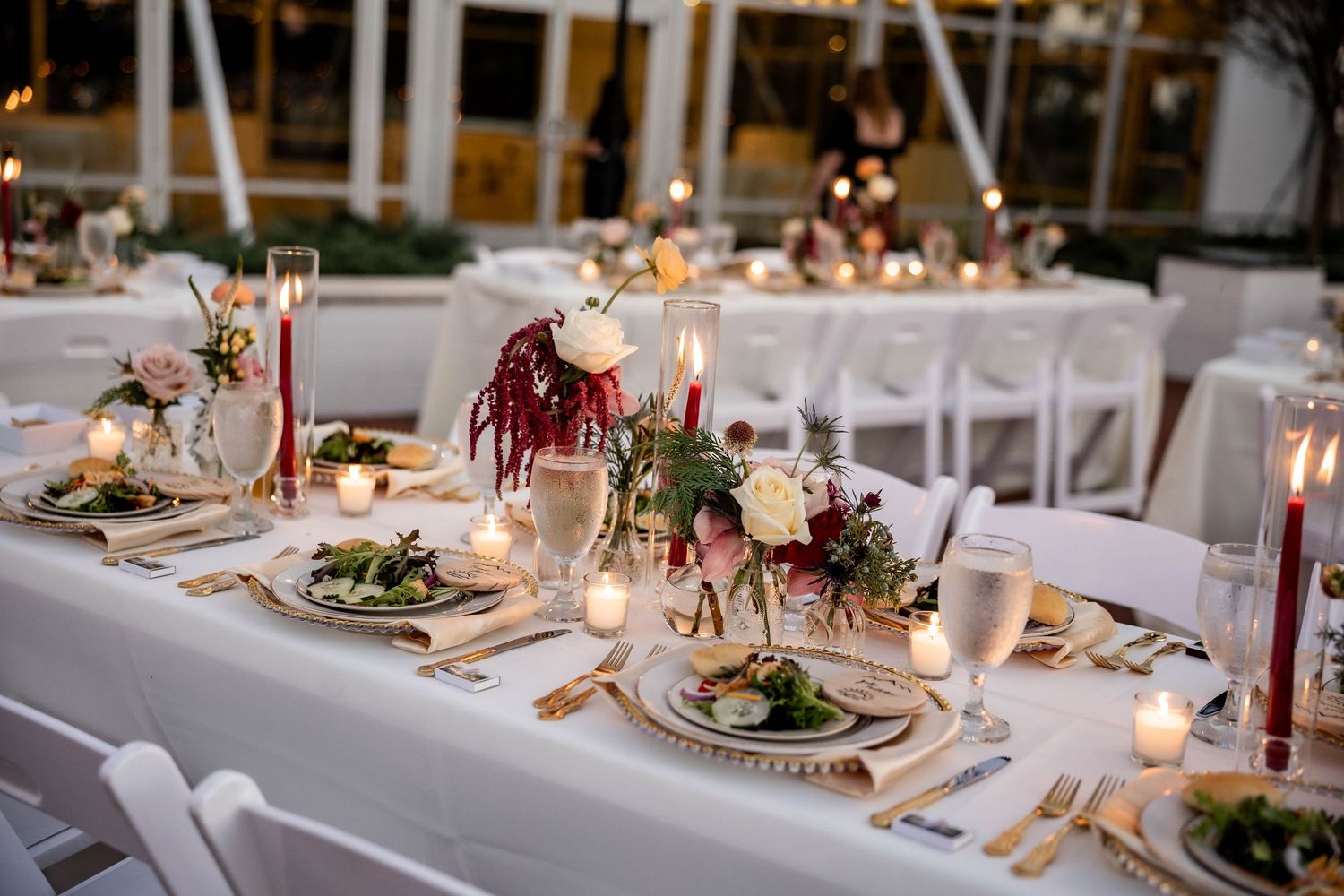 Elegant wedding reception table setting with burgundy floral centerpieces, candles and gold charger plates.