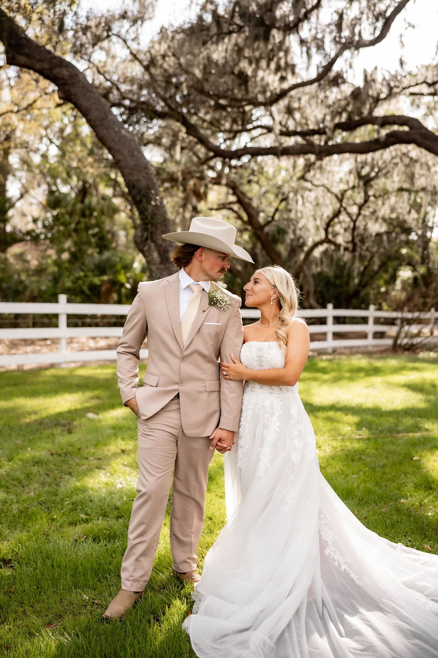A bride and groom share a romantic moment outdoors, dressed in a white lace gown and tan suit with cowboy hat.