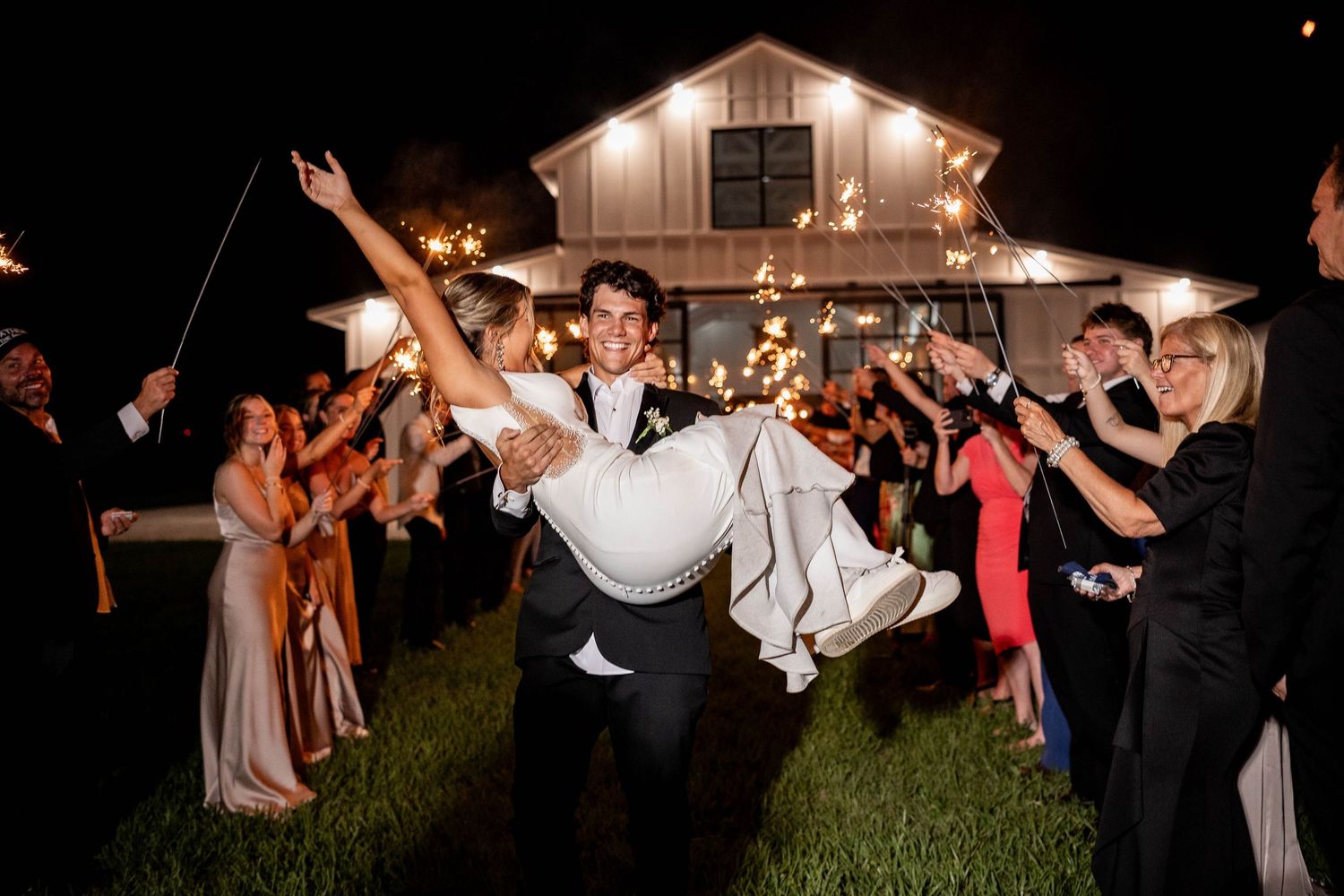 A group of wedding guests toss a person in the air during a nighttime celebration outside.