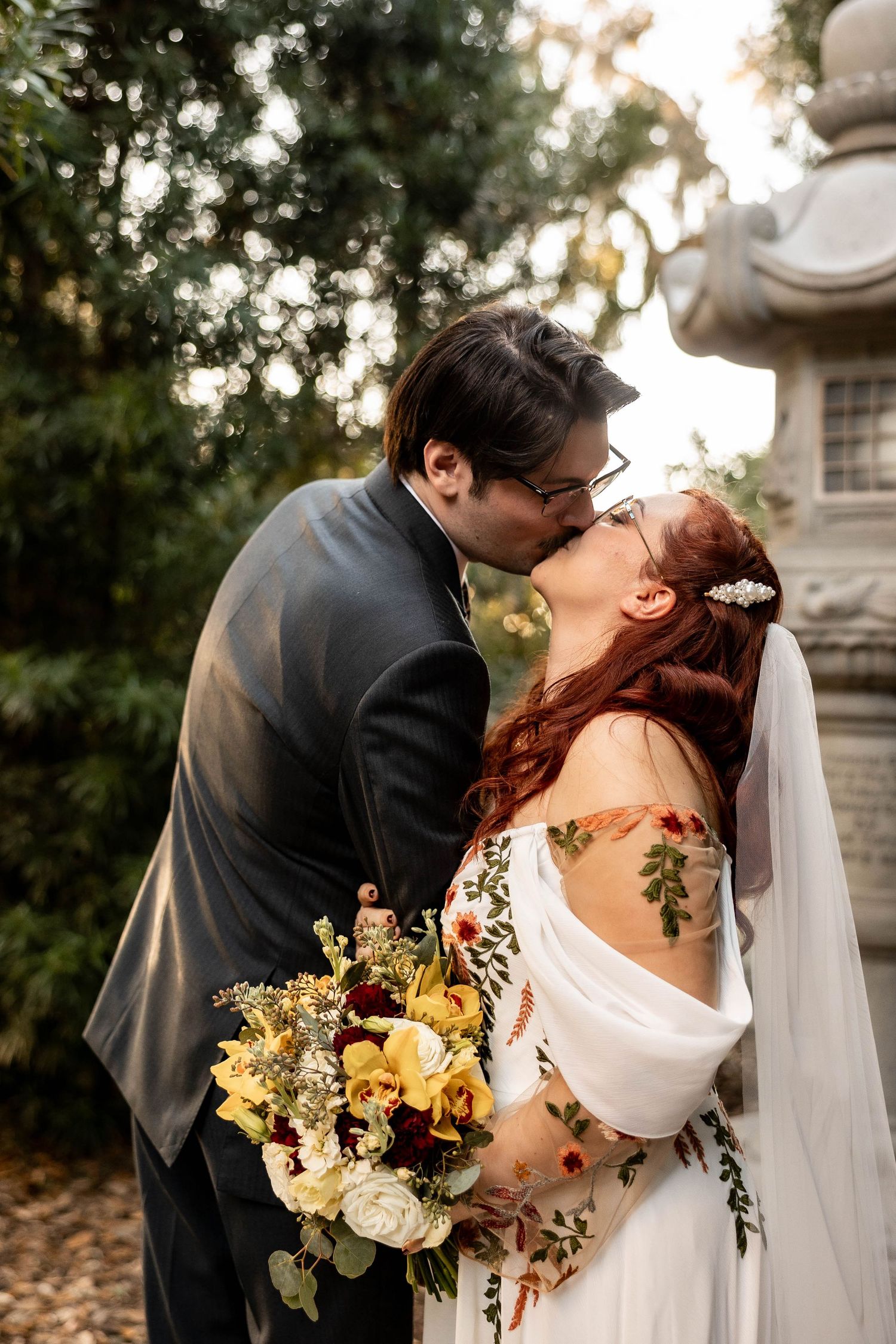 Bride and groom sharing a romantic kiss outdoors, bride holding yellow floral bouquet wearing embroidered off-shoulder wedding dress.