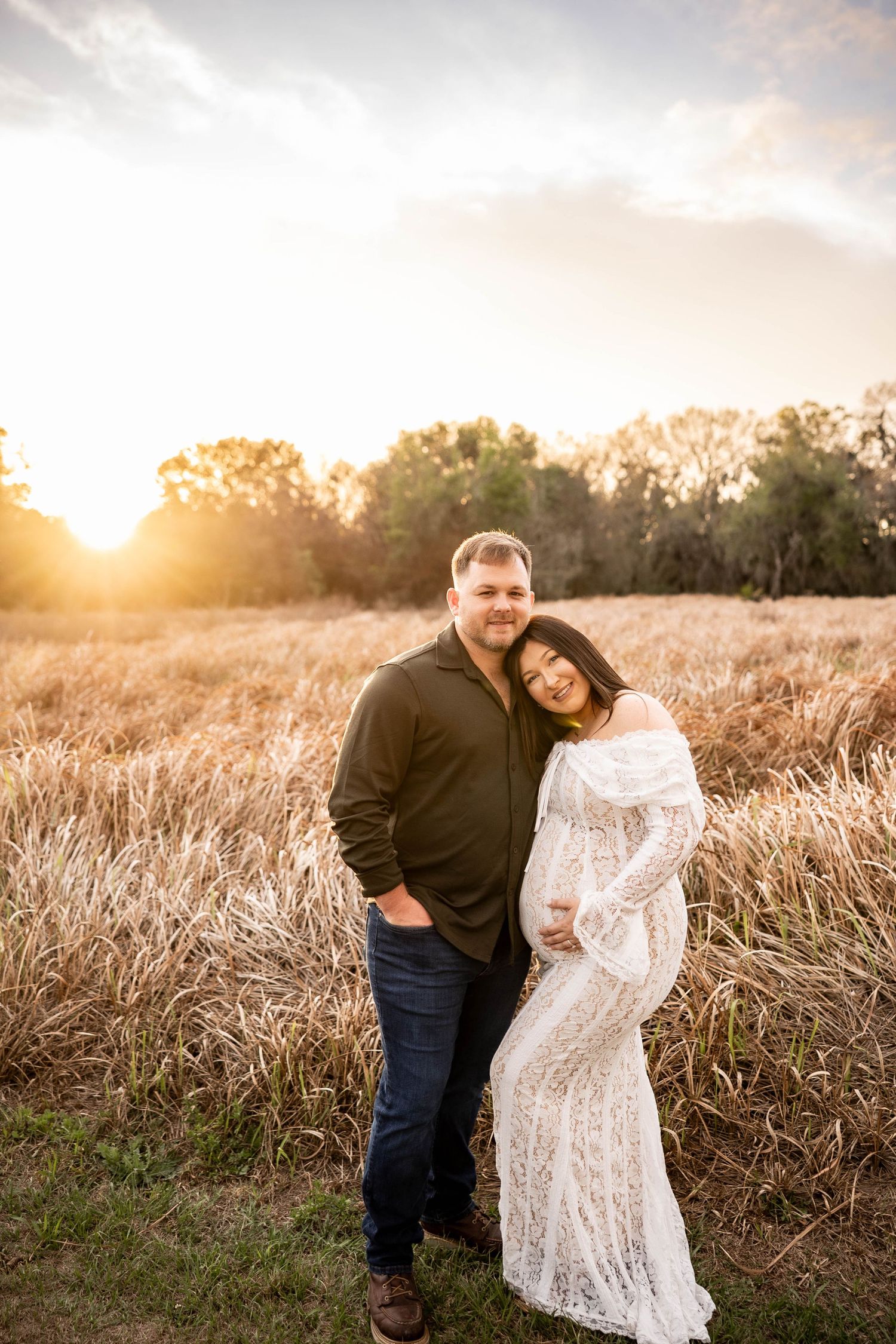 Couple posing at sunset in a golden field, woman in white lace maternity gown leaning on man in dark shirt.