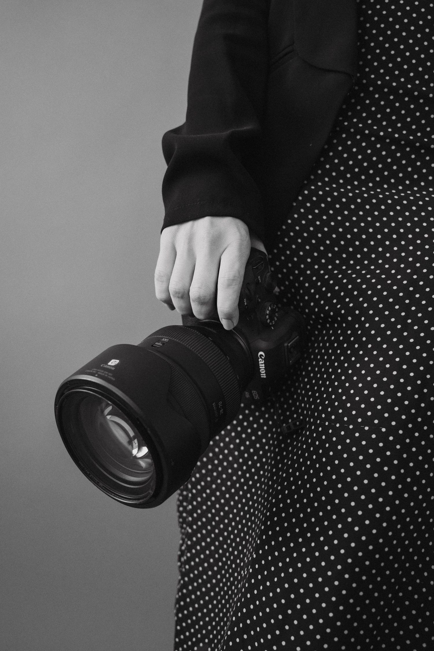 A hand holding a black professional camera lens against a polka dot patterned dress in black and white.
