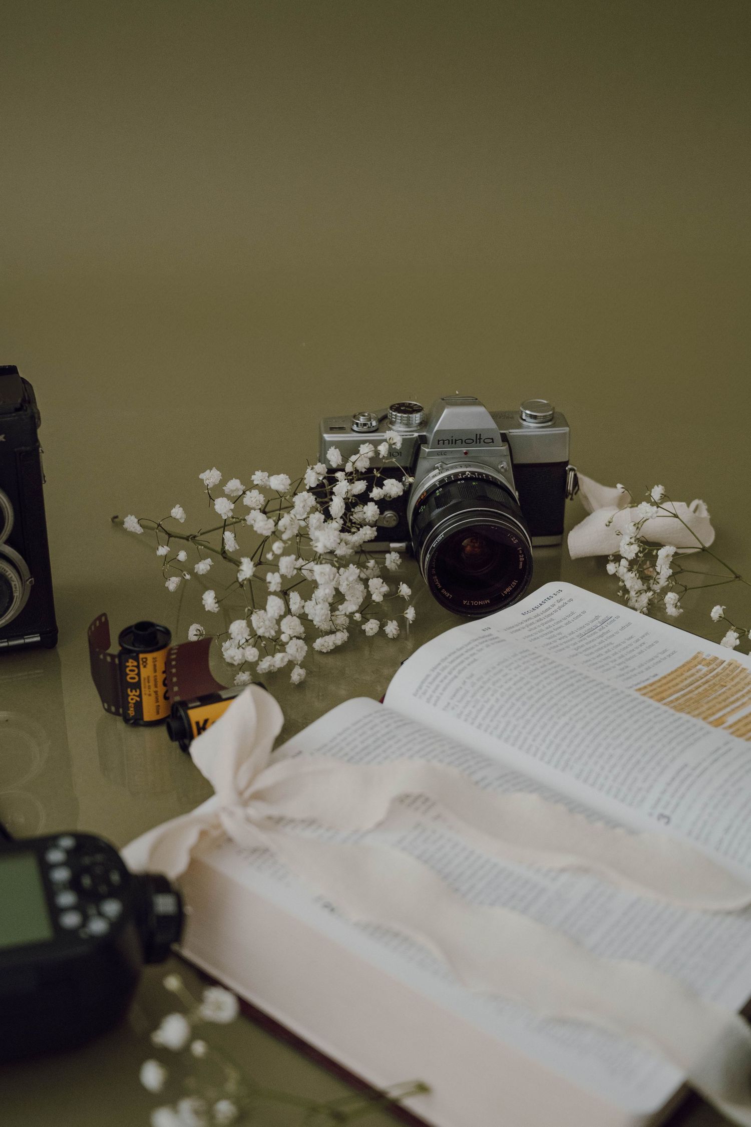 A vintage film camera and roll of film next to an open book decorated with white baby's breath flowers on a green surface.