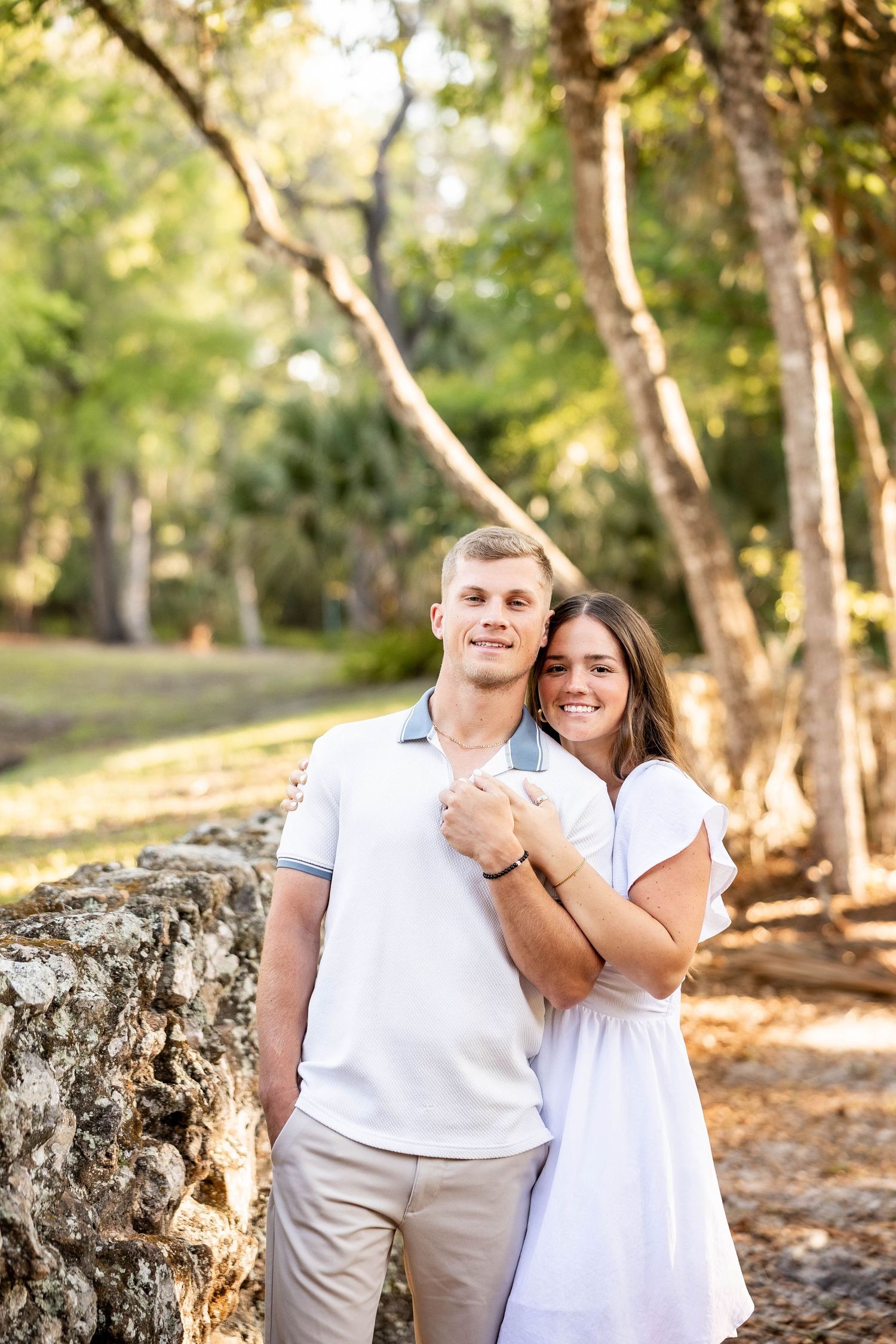 A smiling couple poses outdoors near a stone wall, surrounded by lush green trees, wearing white outfits.